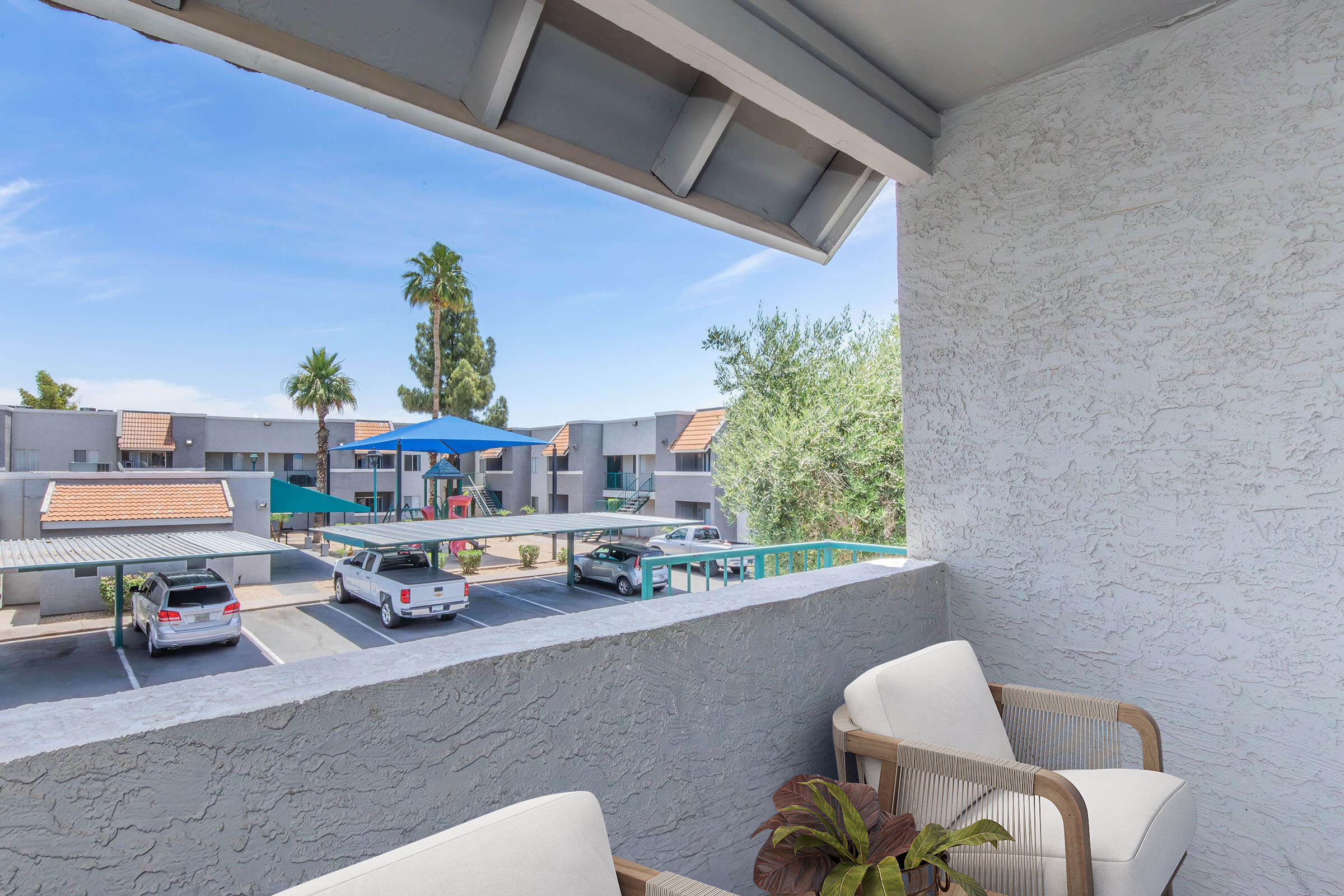 View from a balcony overlooking a parking area and a community pool surrounded by palm trees and apartment buildings. Two chairs are placed on the balcony, with a potted plant nearby. The sky is clear and blue, creating a pleasant outdoor atmosphere.