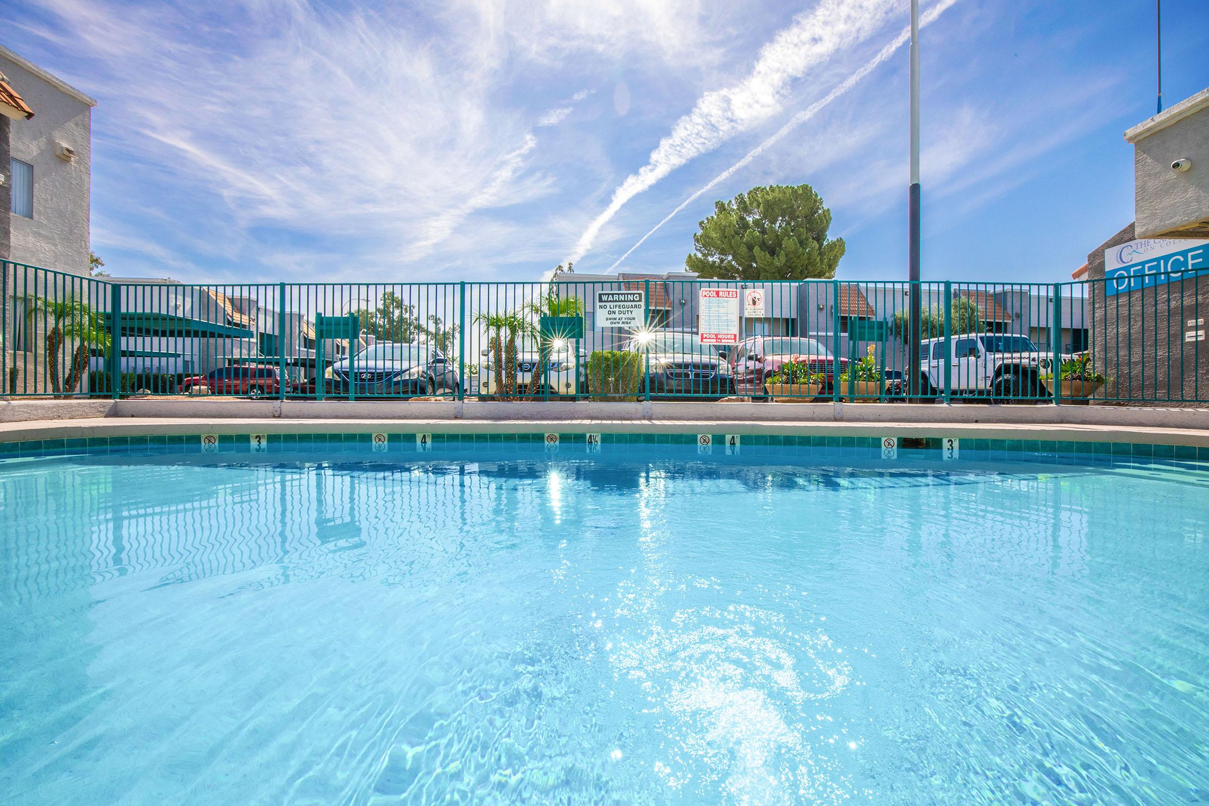 Clear blue swimming pool with a reflective surface, surrounded by green fencing and palm trees. In the background, there are buildings and a clear blue sky with wispy clouds and contrails. The scene conveys a relaxing atmosphere in a sunny environment.