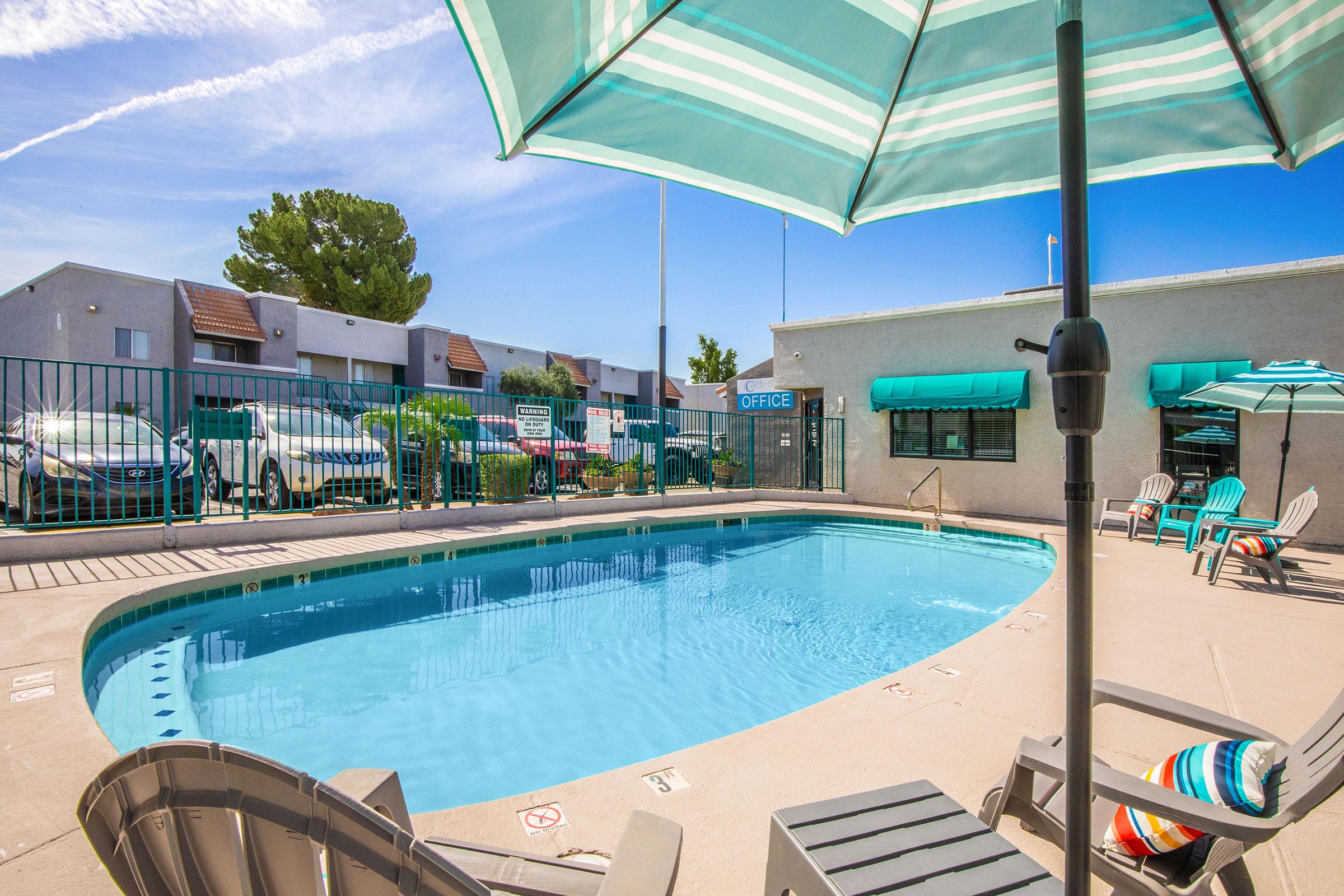 A swimming pool surrounded by lounge chairs and umbrellas, with an office building in the background. The scene is sunny, featuring a clear blue sky and green trees, creating a welcoming atmosphere for relaxation and leisure.