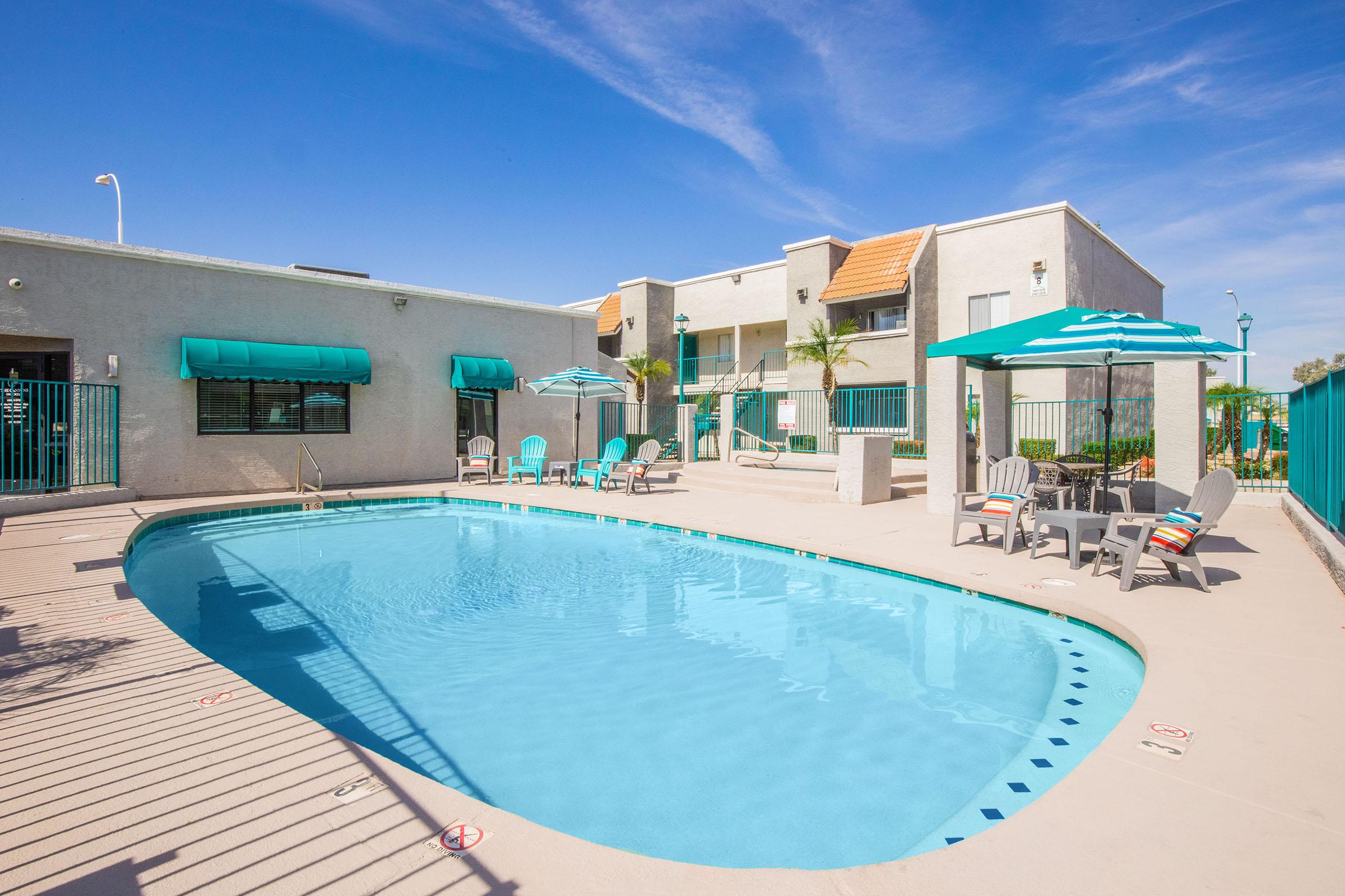 A clear blue swimming pool surrounded by lounge chairs and umbrellas at a hotel. The pool area features palm trees and is enclosed by a fence. The sky is bright and sunny, enhancing the inviting atmosphere of the outdoor space.