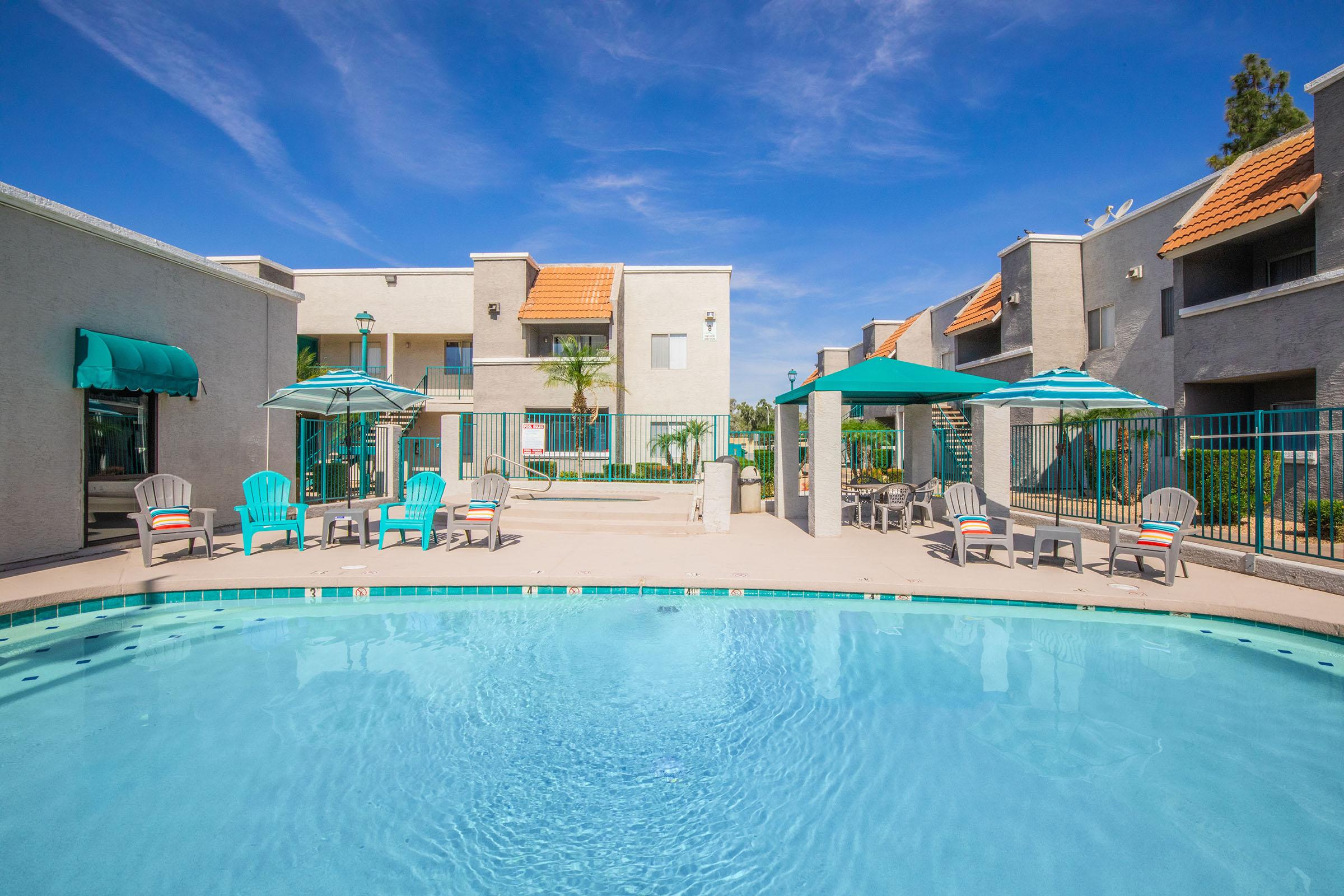 A sunny pool area featuring a clear blue pool surrounded by lounge chairs and umbrellas. Surrounding the pool are modern apartment buildings with palm trees and blue skies in the background, creating a relaxing and inviting atmosphere.