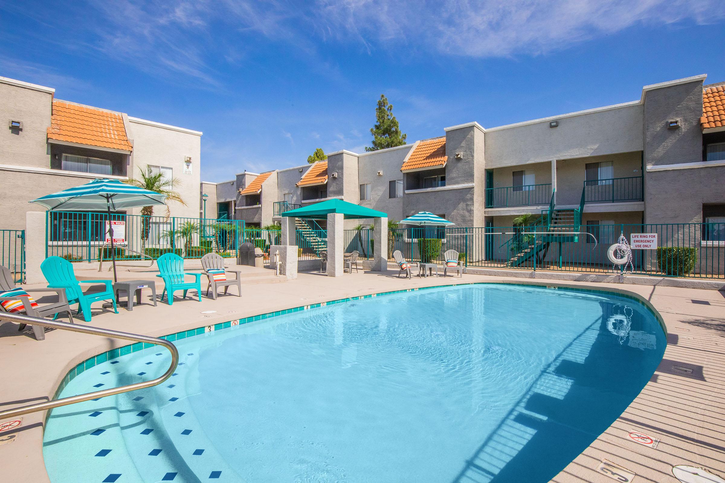 A swimming pool surrounded by lounge chairs and umbrellas, with two-story apartment buildings in the background. The scene is set against a clear blue sky, with palm trees adding a tropical touch. The pool area is well-maintained, creating a welcoming atmosphere for relaxation and leisure.