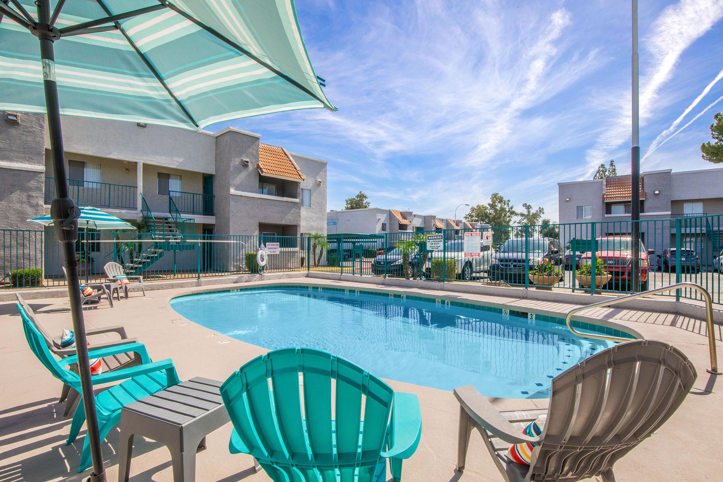 A tranquil pool area featuring a clear blue swimming pool, surrounded by turquoise and gray lounge chairs. An umbrella provides shade, with an apartment complex in the background. The sky is bright and partly cloudy, creating a relaxing outdoor atmosphere.