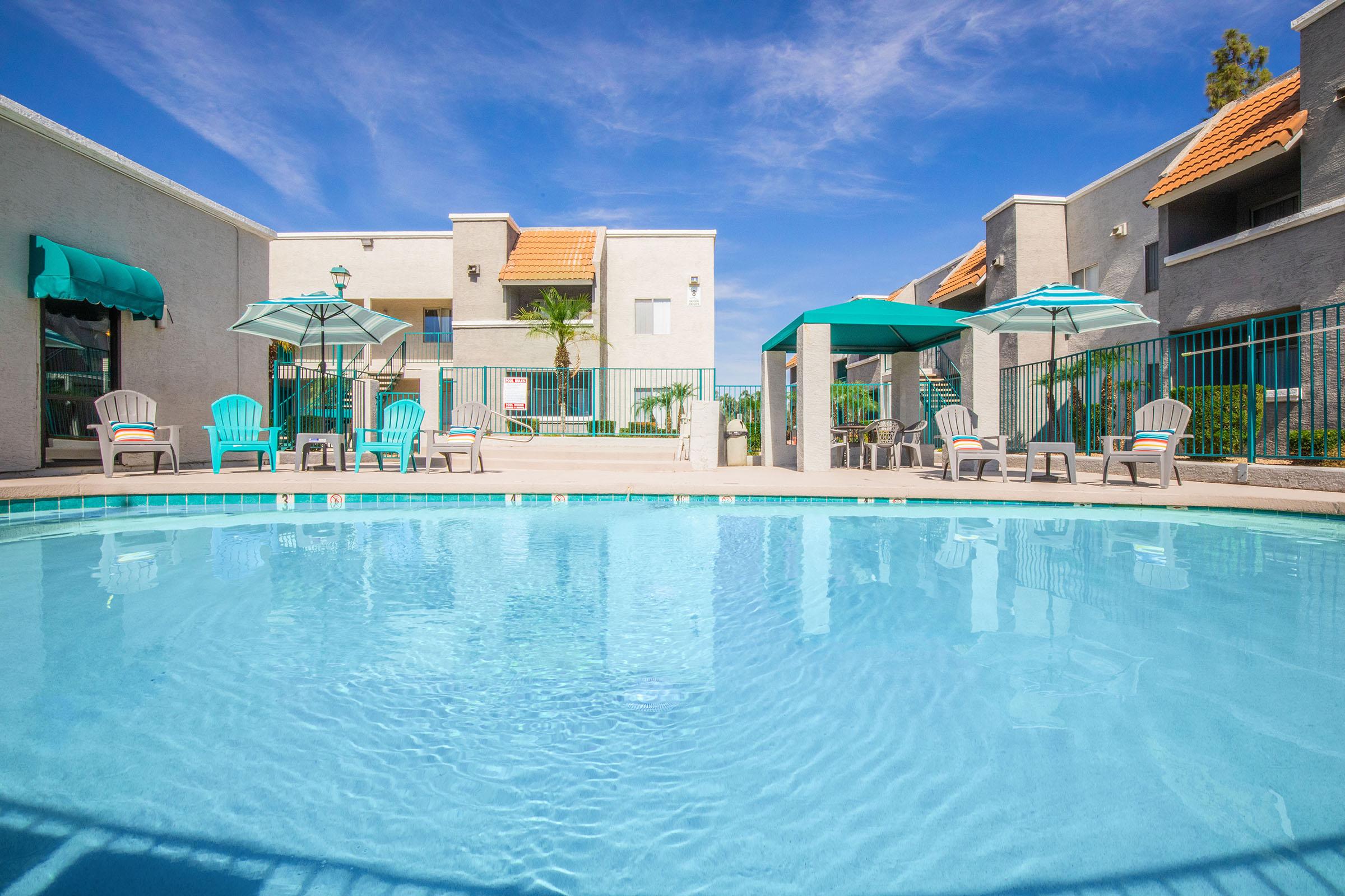 A clear swimming pool surrounded by lounge chairs and umbrellas, with residential buildings in the background. The sky is blue with few clouds, creating a bright and inviting atmosphere for relaxation and leisure.