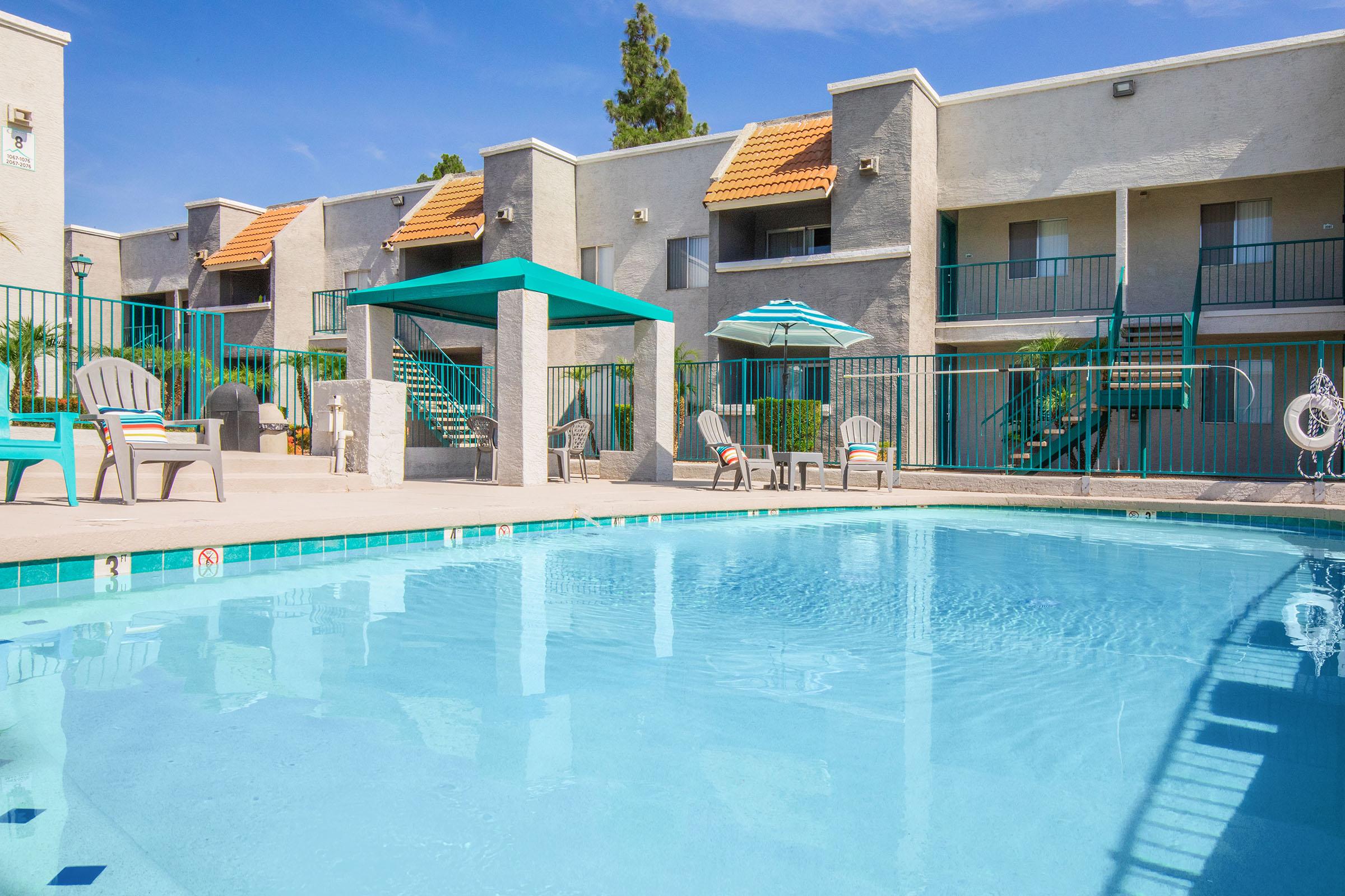 Swimming pool in a residential complex, featuring clear blue water, lounge chairs, a shaded gazebo, and an umbrella. The background showcases a two-story building with several balconies and greenery, under a bright blue sky.