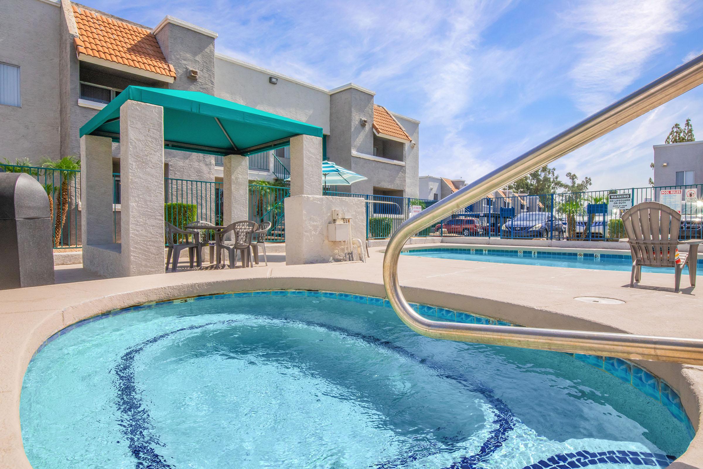 A serene outdoor scene featuring a sparkling hot tub and swimming pool area, with a shaded gazebo and patio seating nearby. The sky is bright blue with wispy clouds, and there are several lounge chairs arranged by the pool, creating a relaxing atmosphere for leisure.