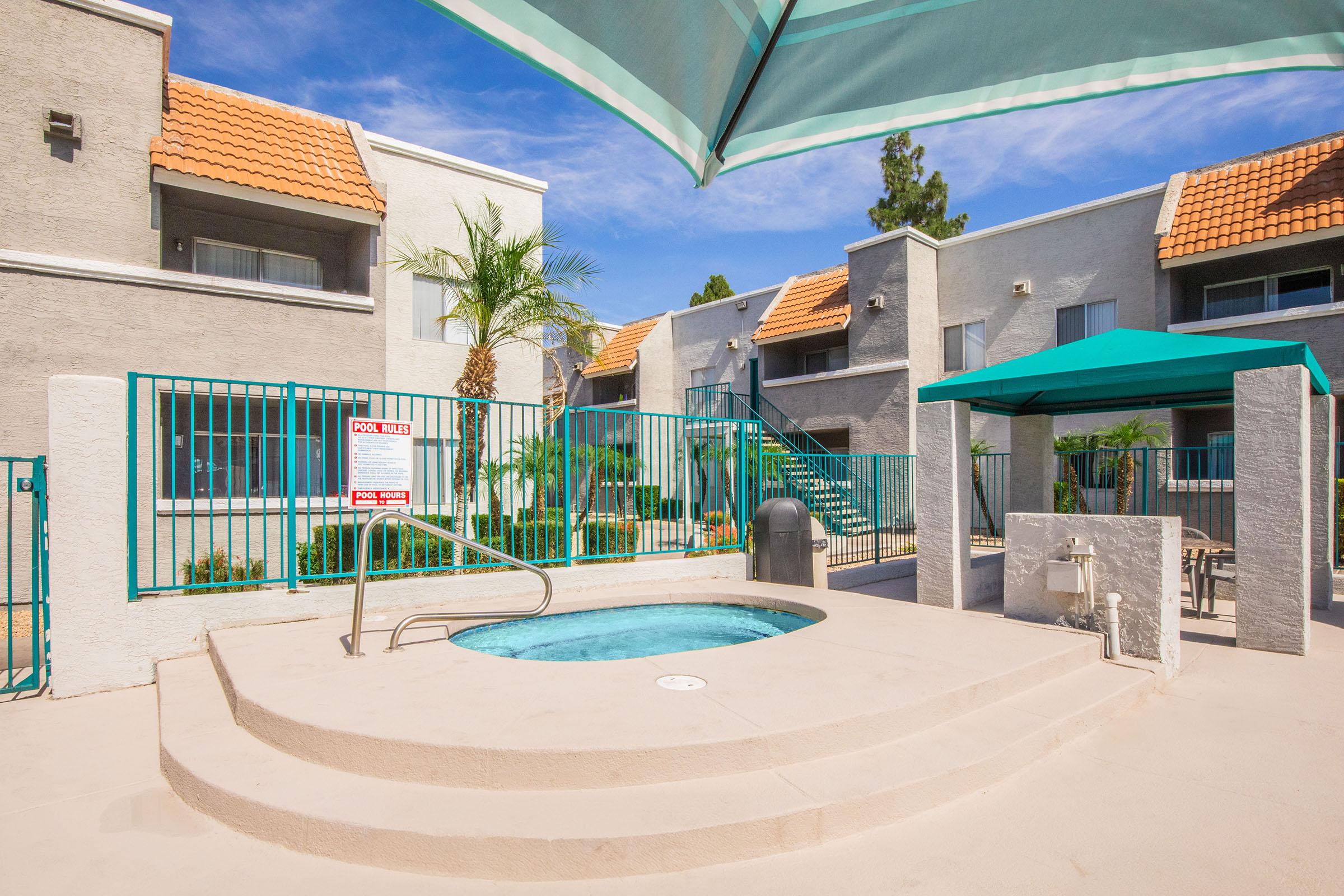 A sunny outdoor space featuring a hot tub surrounded by a gated area and palm trees, with a couple of residential buildings in the background. There is a green umbrella over a seating area, and a sign indicating parking restrictions. The sky is clear and blue, enhancing the tranquil atmosphere.