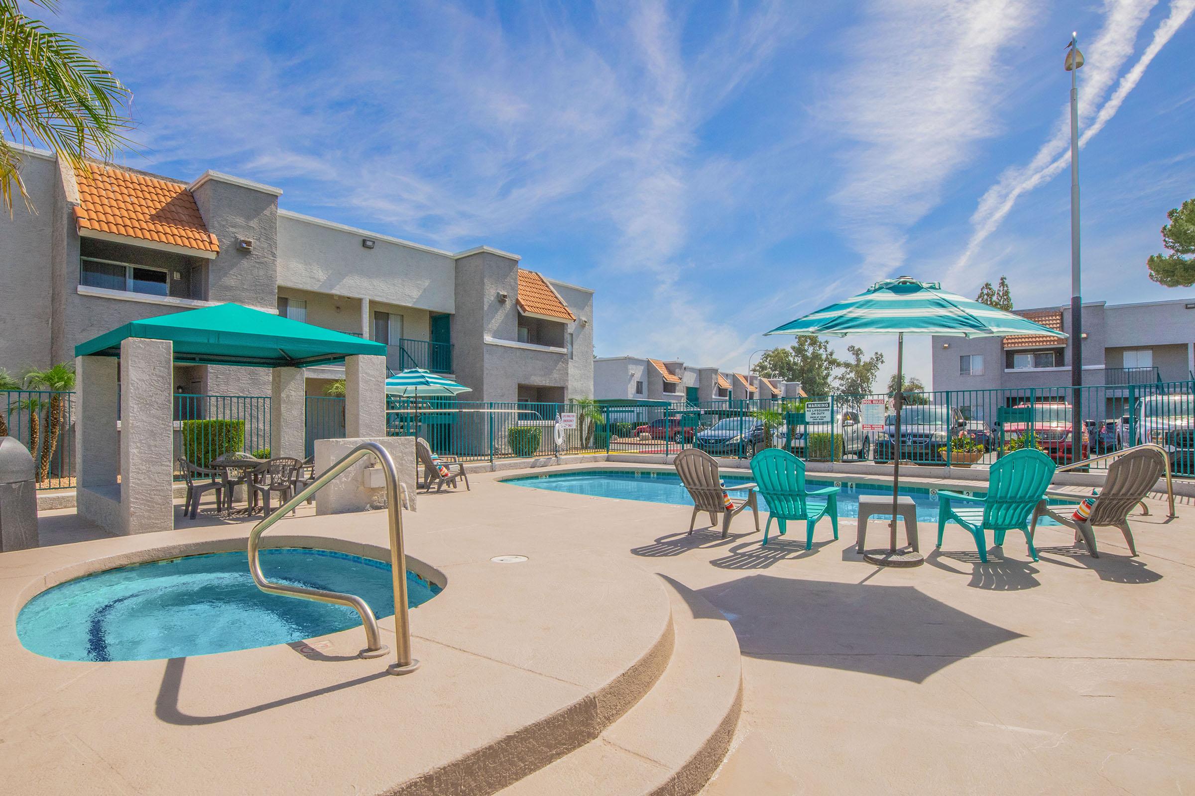 A sunny outdoor pool area featuring a large swimming pool and a hot tub. There are several turquoise lounge chairs and umbrellas, with palm trees in the background. Residential buildings are visible behind the pool area, under a bright blue sky with wispy clouds.