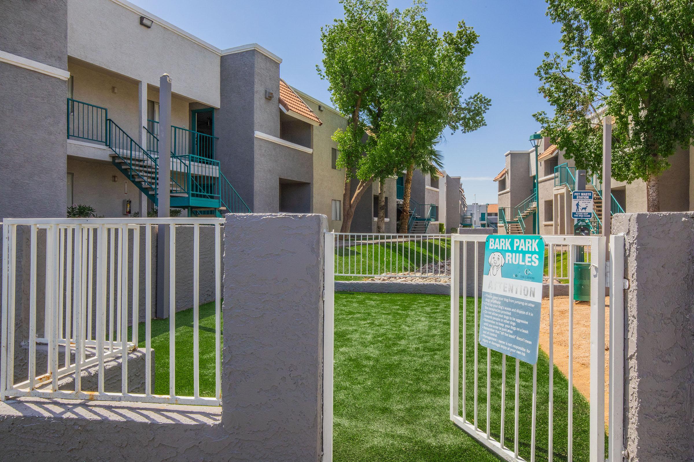 View of an apartment complex featuring a gated entrance to a dog park area. The park has green artificial grass, signs displaying park rules, and surrounding apartment buildings with trees and pathways visible. Bright blue sky above adds to the inviting atmosphere.