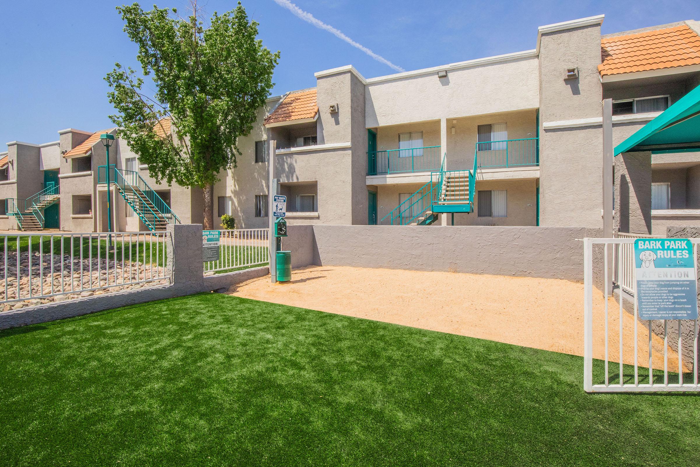 A view of a pet-friendly apartment complex featuring two-story buildings with balconies, green staircases, and a fenced dog park with artificial turf. The park includes a sign displaying rules for pet owners. The area is sunny with clear blue skies and lush trees in the foreground.