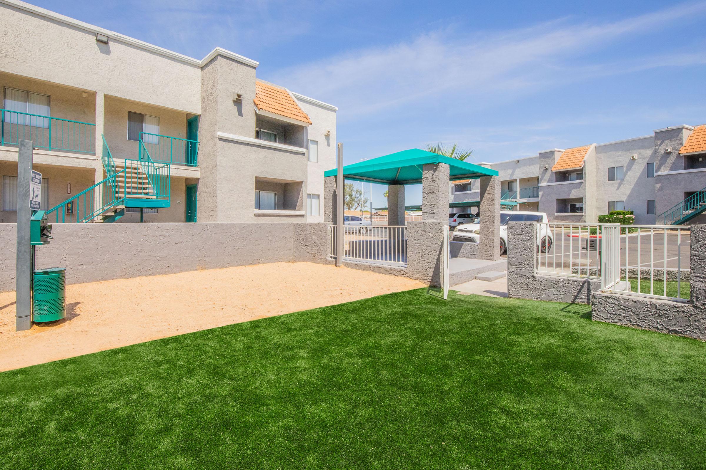 View of a landscaped area featuring artificial grass and a sandy section. In the background, there's a two-story apartment complex with light-colored walls and orange roof tiles. A green shade structure is visible nearby, along with a fenced area and parking. The sky is clear and blue, providing a bright, sunny atmosphere.
