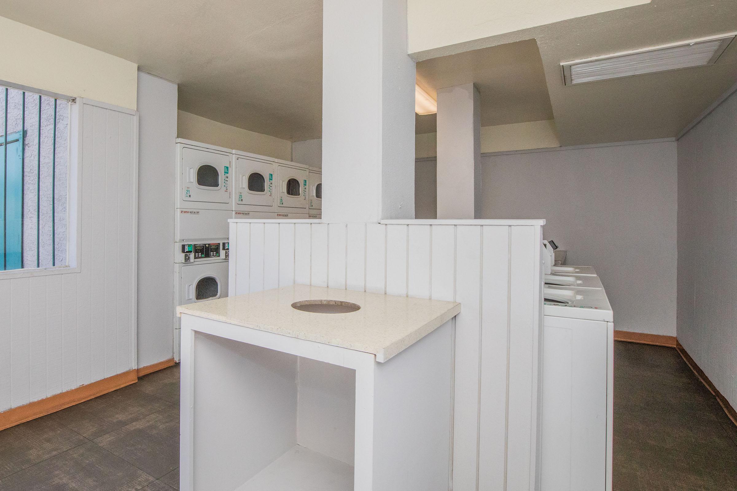 A clean and well-lit laundry room featuring several stacked dryers and washing machines along the wall. There's a white counter with a circular opening in the center, and the walls are painted in light colors. A window with bars provides natural light. The floor is tiled with gray tiles.