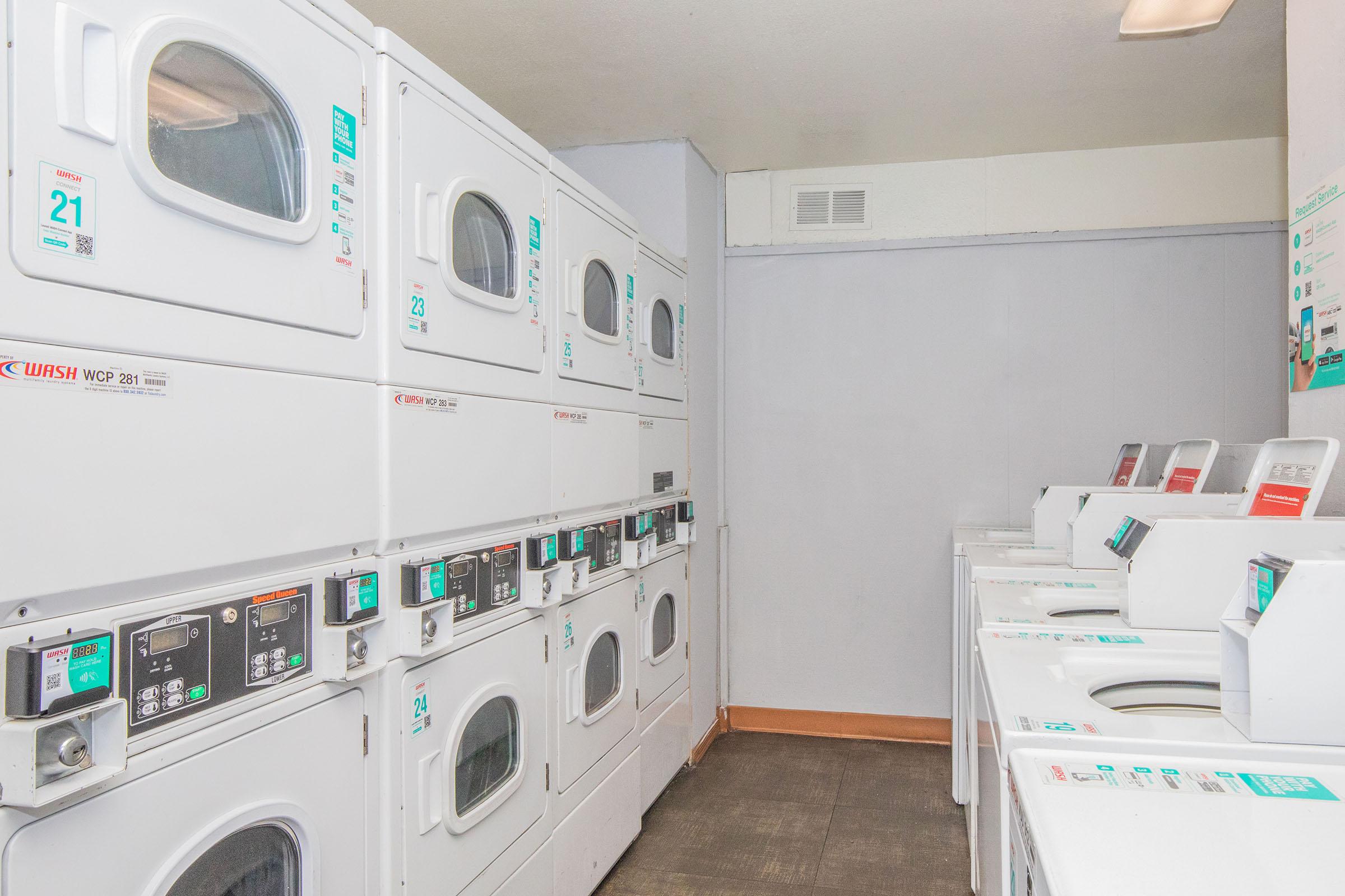A clean and tidy laundry room featuring multiple stacked washers and dryers. The machines display digital controls and are arranged against a light-colored wall, with a neutral floor. The space appears organized and ready for use, providing a convenient setting for laundry tasks.
