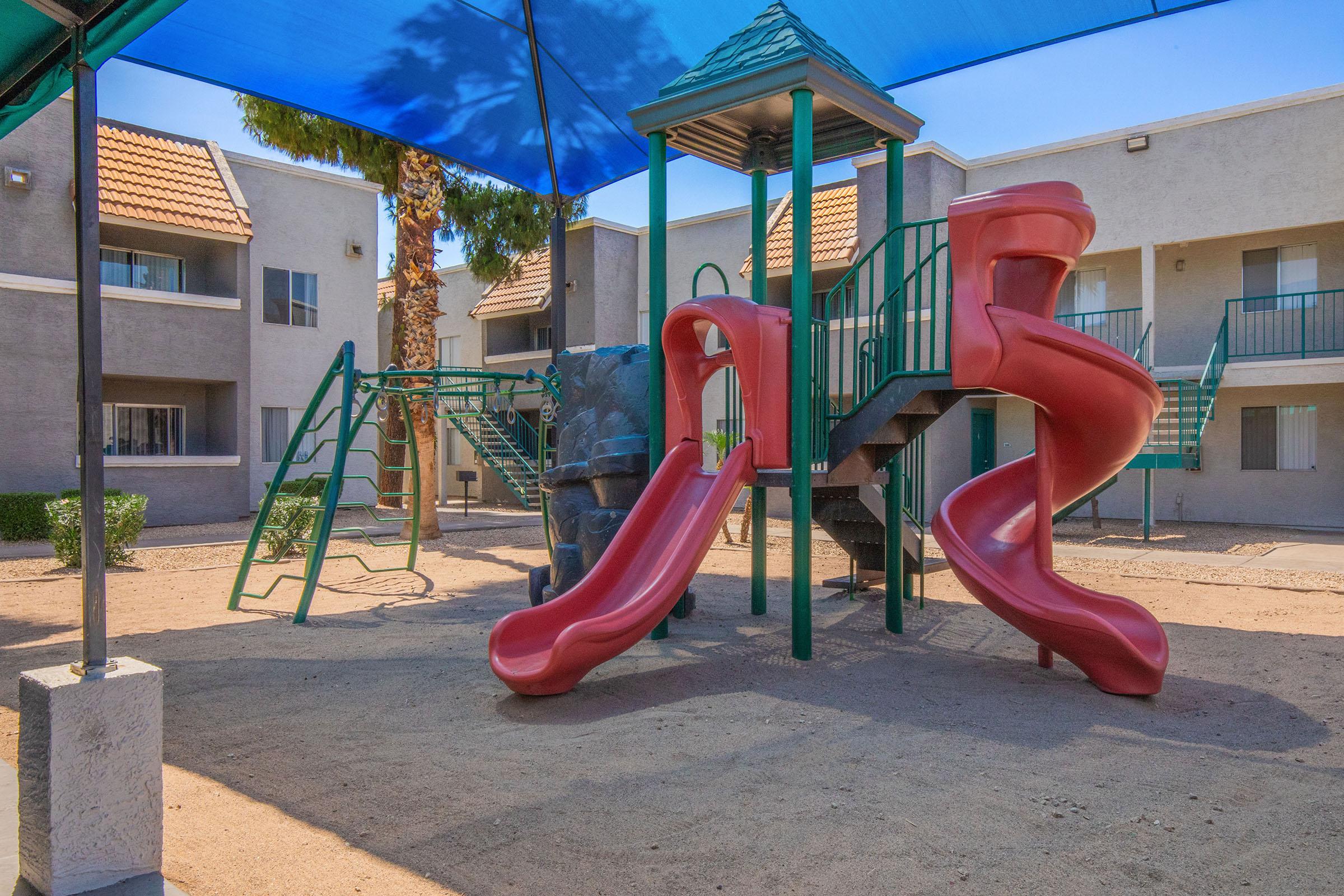 A playground featuring two red slides, a climbing structure, and a green swing set, all situated under a blue shade canopy. The background includes gray apartment buildings with balconies and palm trees. The ground is sandy, providing a casual play environment.