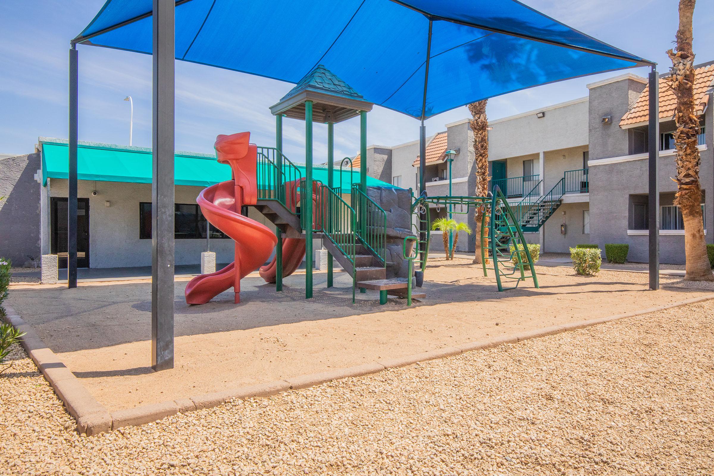 Playground area featuring a red slide, green climbing structure, and swings, all shaded by a blue canopy. Surrounding the playground are palm trees and a gravel surface, with nearby buildings partially visible in the background. The scene is bright and sunny, creating a welcoming outdoor space for children.