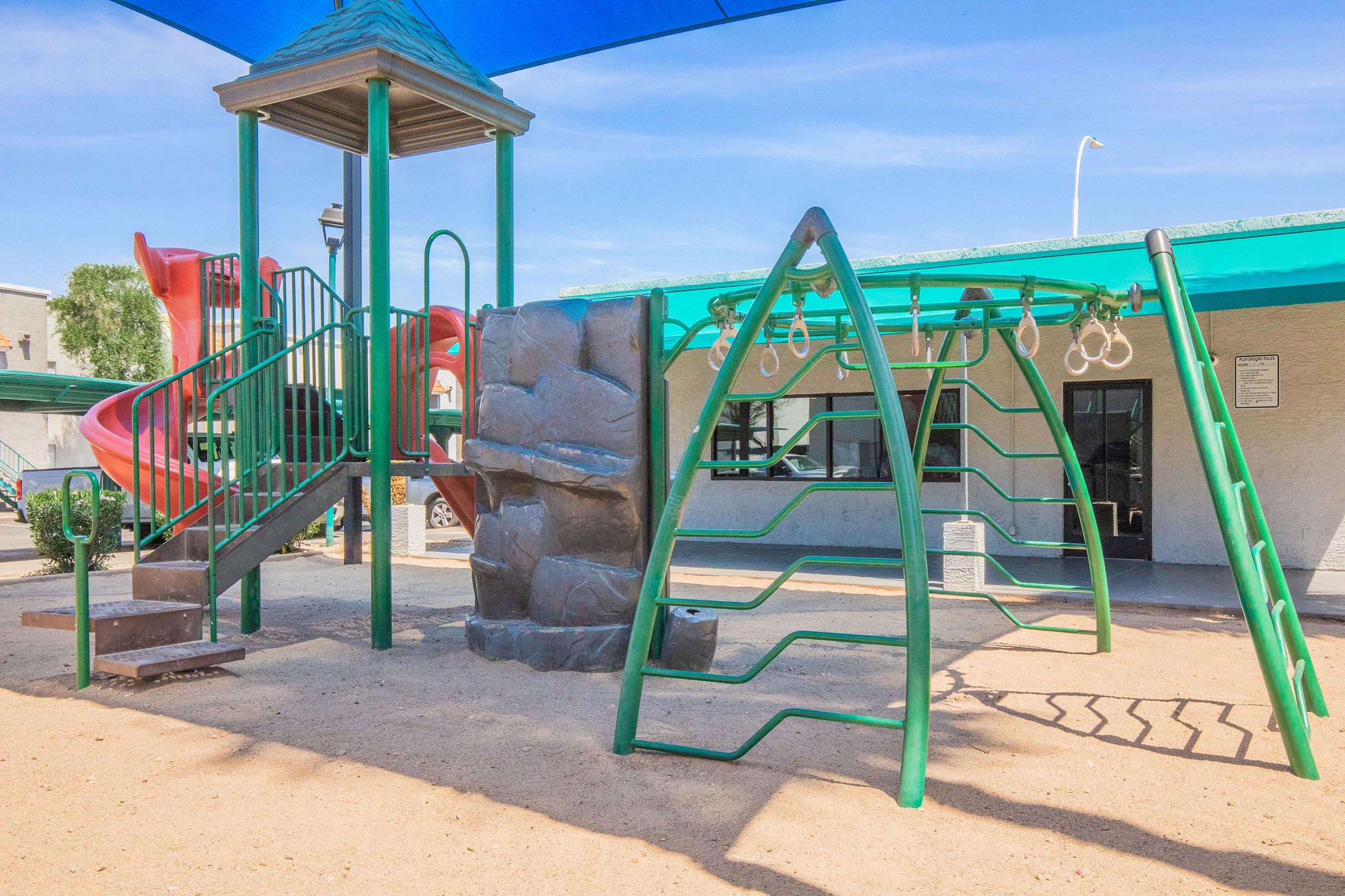 Colorful playground featuring a tall green slide, climbing structure with a rock wall, and a set of monkey bars. A shaded area provides protection from the sun, with sand below and a clear blue sky in the background. Suitable for children's play and outdoor activities.