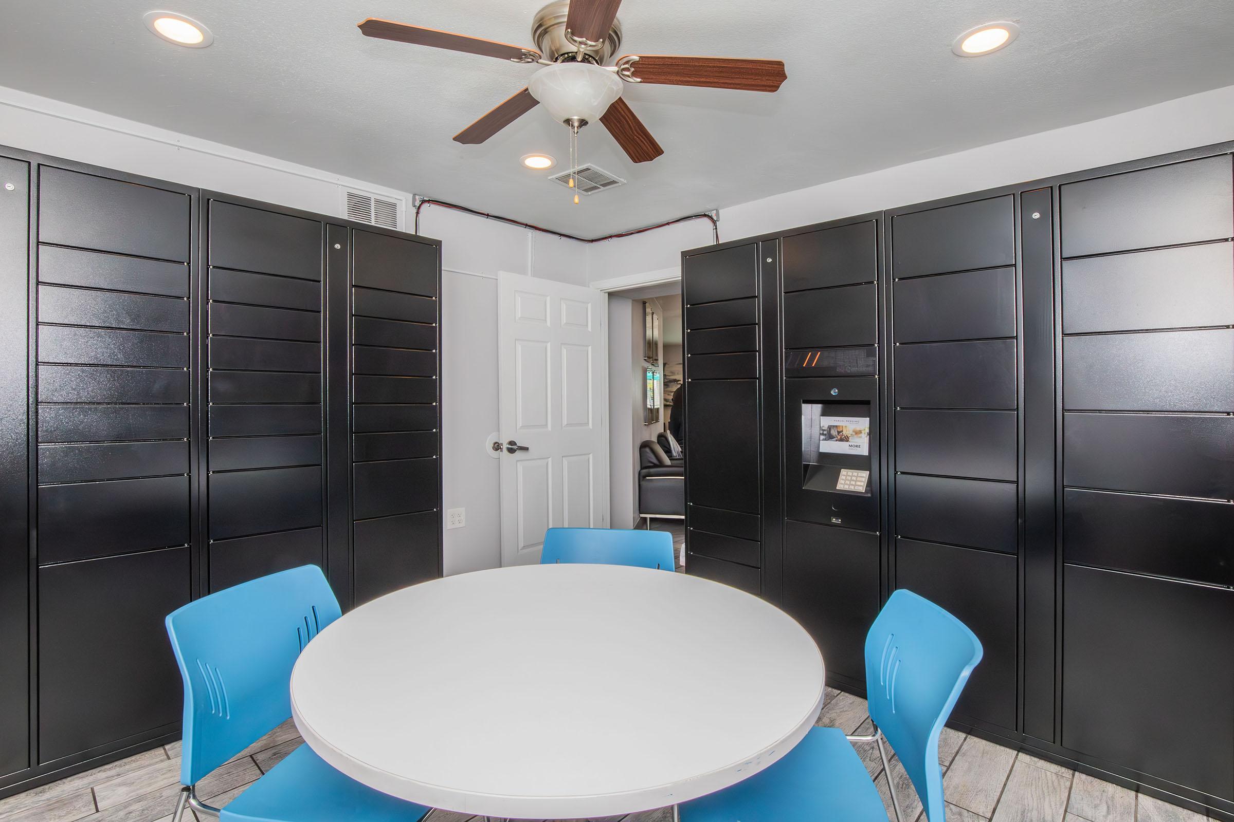 A modern room featuring a round white table surrounded by blue chairs. Black locker units line the walls, and a ceiling fan is visible. A door is partially open, suggesting access to another space, creating a functional and contemporary atmosphere.