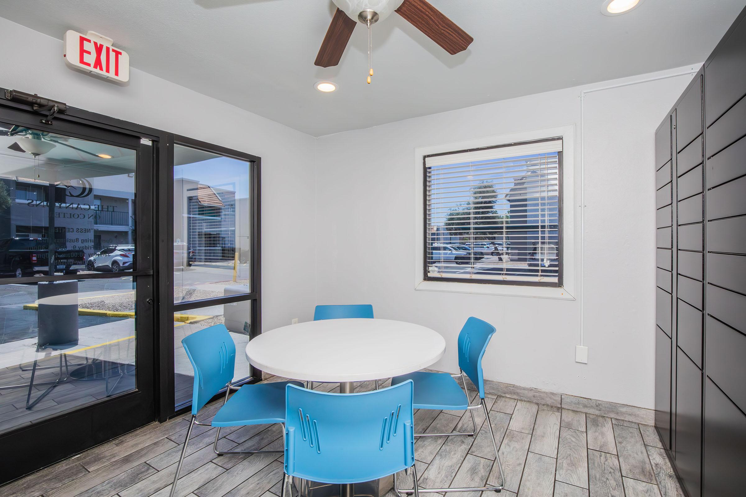 A bright, modern room featuring a circular white table surrounded by four blue chairs. There’s a ceiling fan above and large windows letting in natural light. An exit sign is visible near the door, and a set of lockers is positioned against the wall. The room has a clean, minimalist aesthetic.