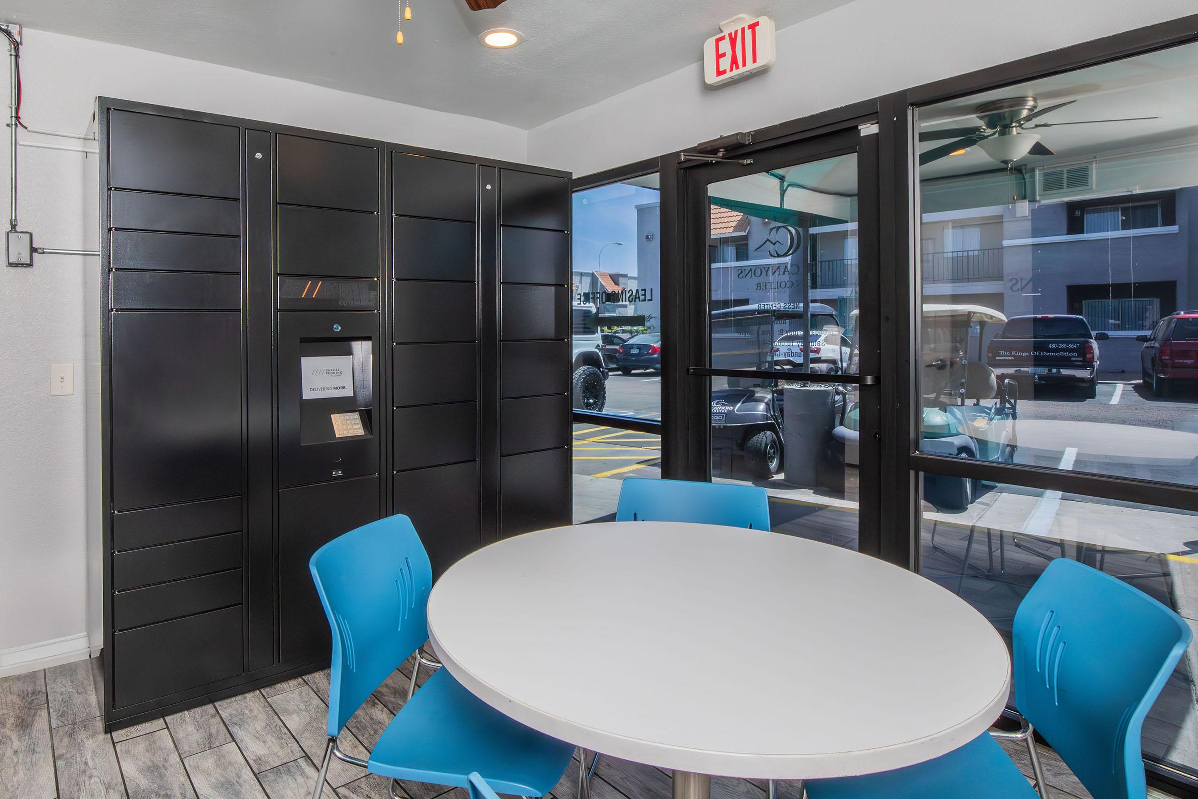 A modern, well-lit interior of a lobby space featuring a black package locker system and a circular white table surrounded by blue chairs. Large glass doors lead outside, with visible parking and vehicles in the background. The setting appears welcoming and functional, ideal for package retrieval.