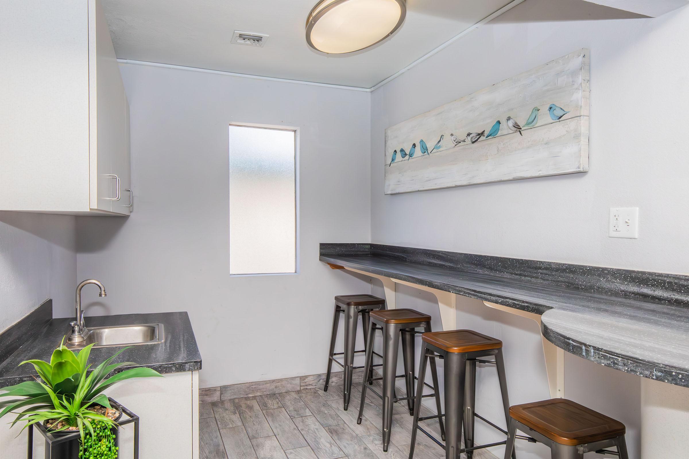 A minimalist kitchen area featuring a gray countertop, a sink, and three wooden stools. A piece of wall art depicting blue birds hangs above the countertop. The walls are painted light gray, and a small window lets in natural light, creating a bright and airy atmosphere. A potted plant adds a touch of greenery.