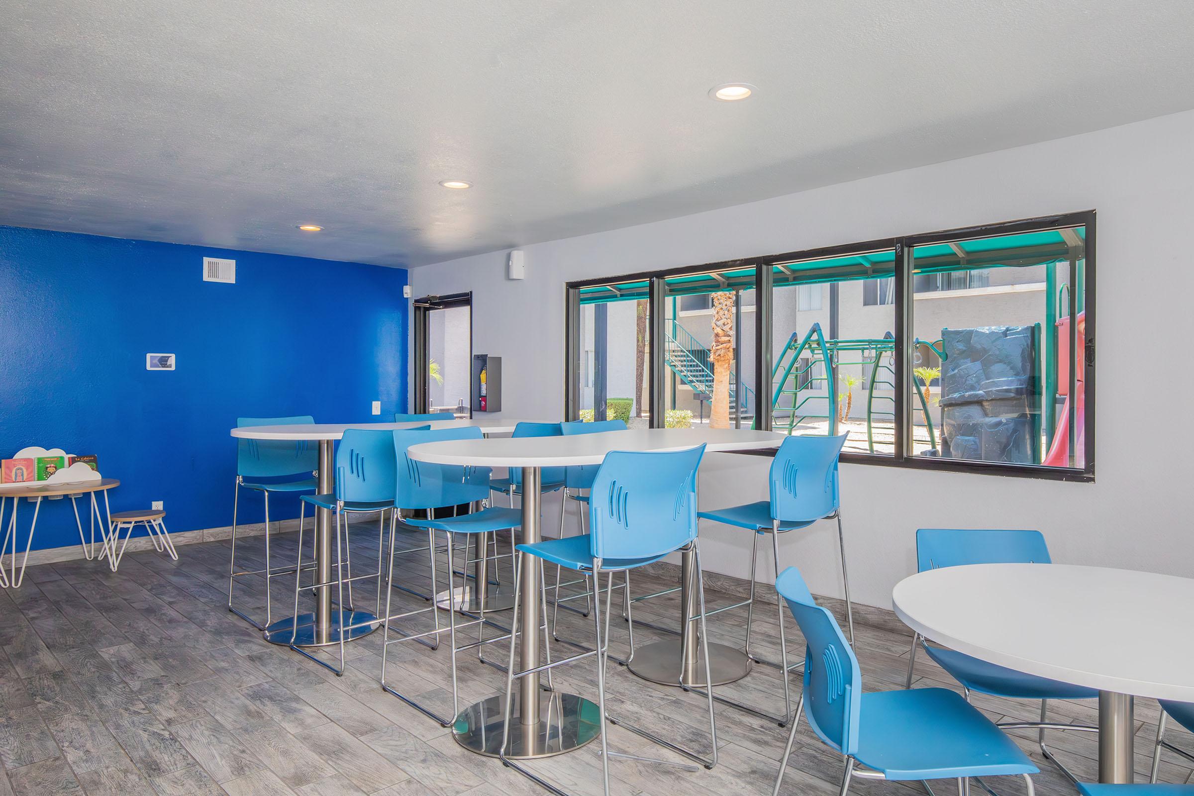 Brightly lit modern room featuring blue and white decor. Tables with white tops and blue chairs are arranged throughout. Large windows provide views of an outdoor area. The flooring is light wood, and the walls are a mix of blue and white. There are playful elements like toys in the corner, adding a lively touch.
