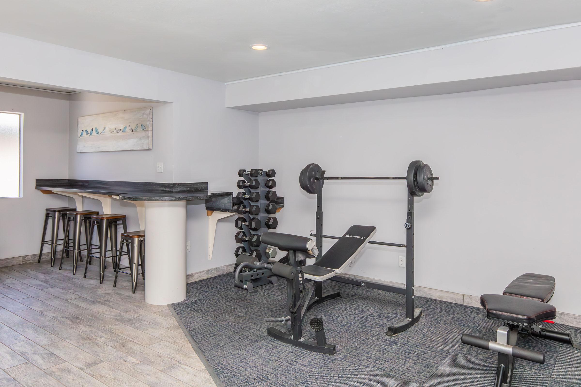 A modern home gym featuring a weight bench with weights, an exercise chair, and a rack of dumbbells. There’s a bar with stools nearby, and the room has neutral-colored walls and tiled flooring, creating a spacious and inviting workout area.