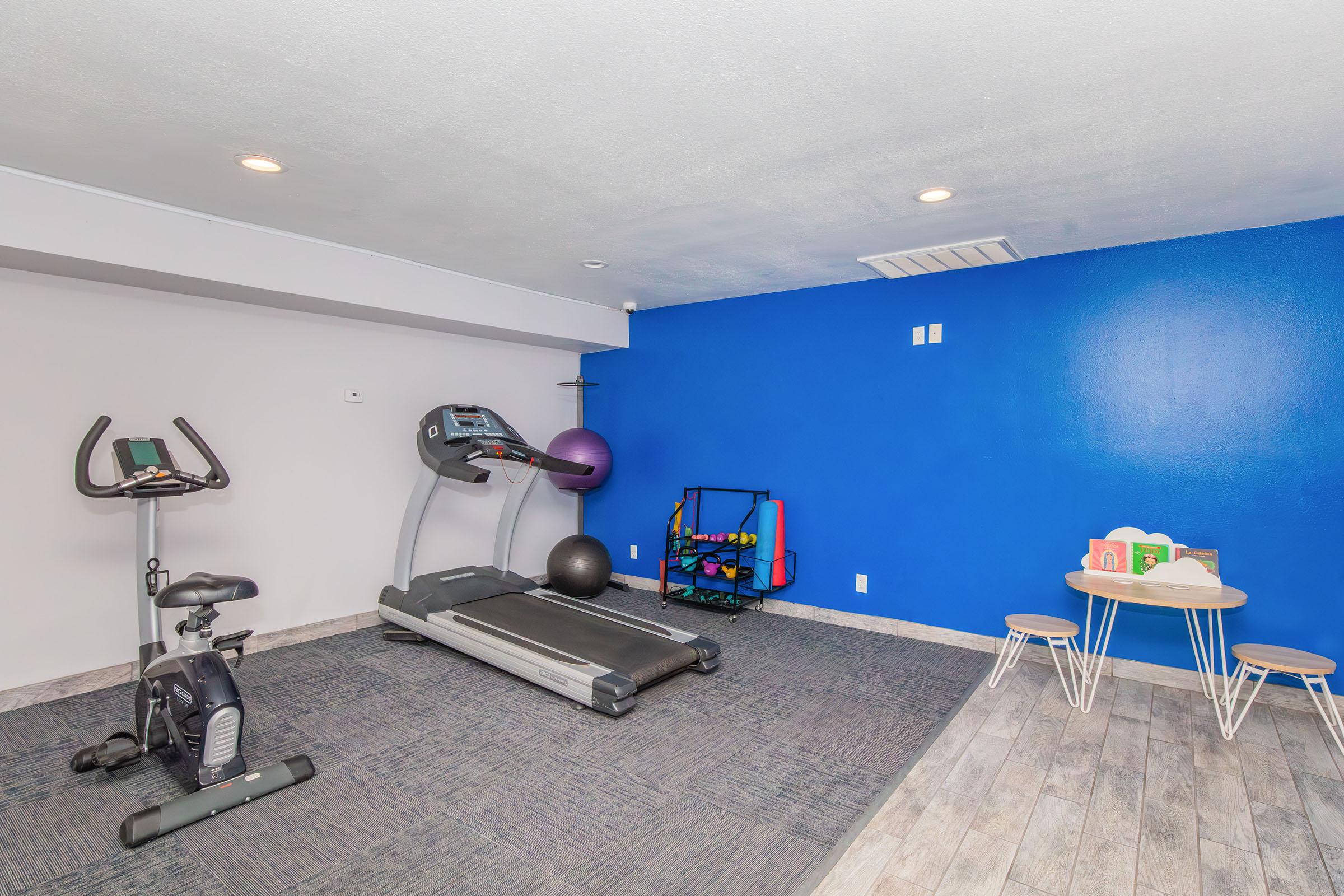 A compact home gym featuring a treadmill, an exercise bike, a stability ball, a rack with weights, and a small table with colorful building blocks against a blue wall, creating an inviting workout space.
