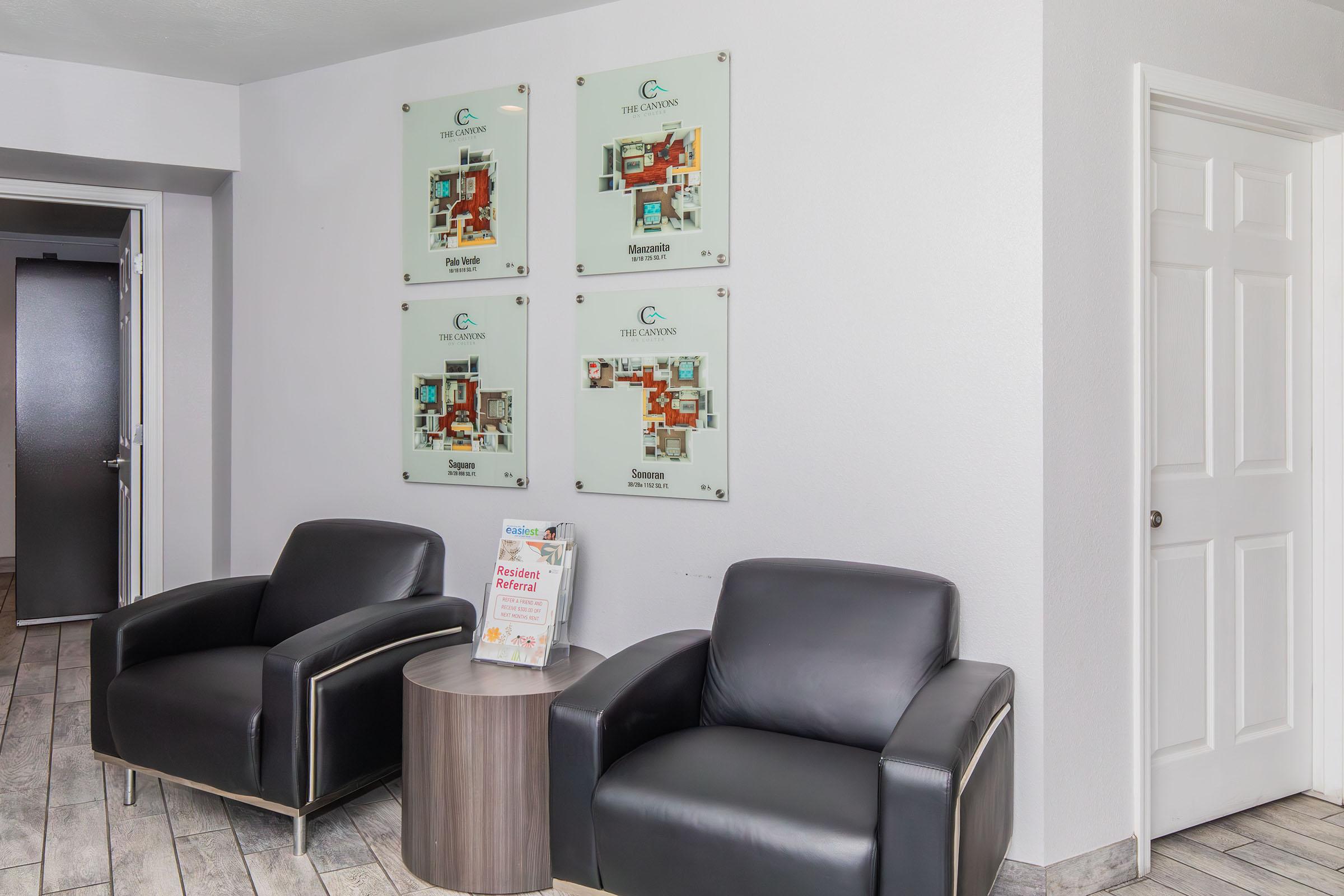 A waiting area featuring two black leather armchairs and a small round table. On the wall are three transparent display boards with floor plans, labeled with names like "The Gardens" and "The Courtyard." A brochure titled "Flexible Referral Program" is placed on the table. The room has a neutral color scheme and modern decor.
