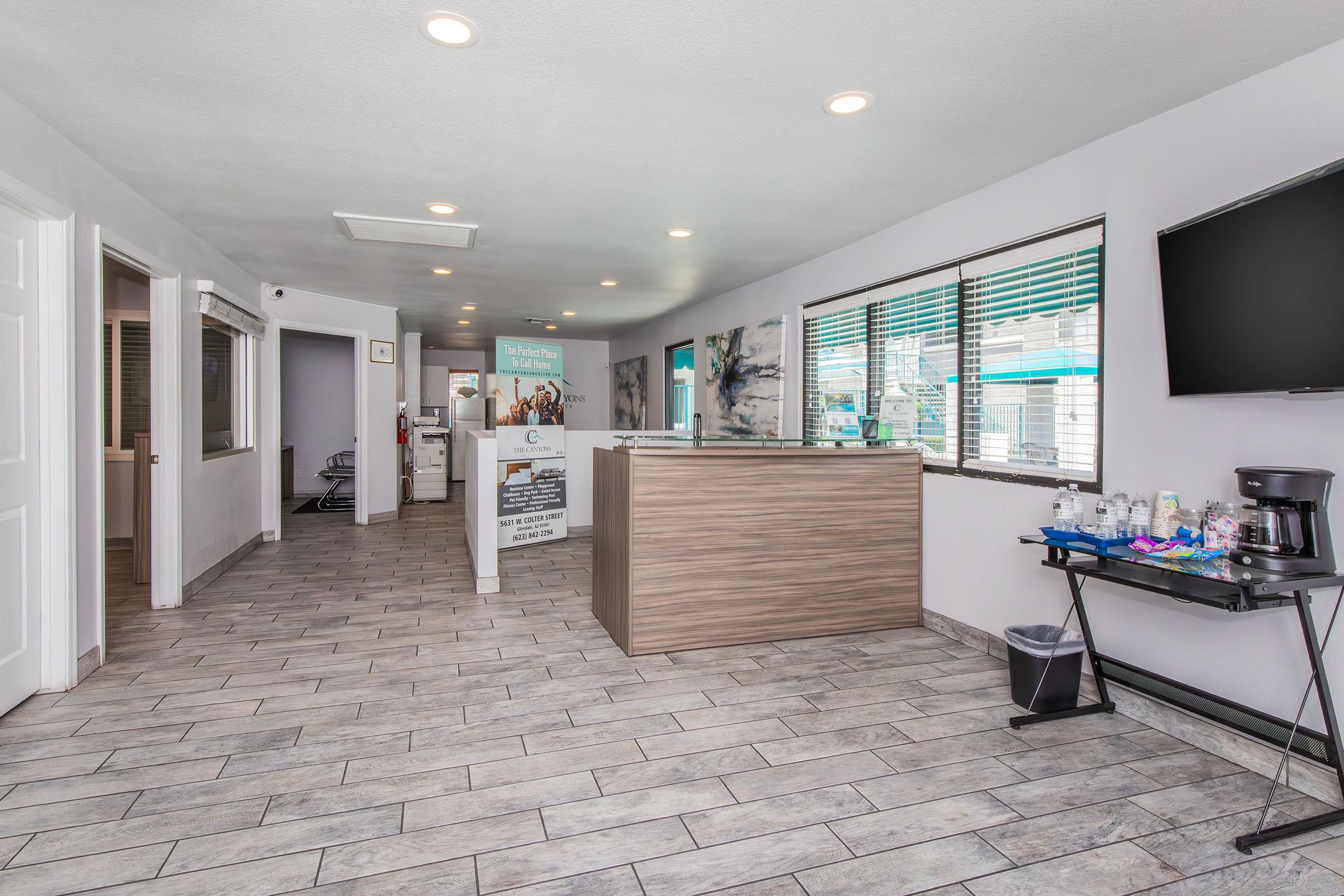 Interior of a modern office reception area featuring a wood-paneled front desk, large windows with natural light, tiled flooring, and a waiting area with a coffee station. The walls are decorated with minimalistic art, and there are informational displays in the background.