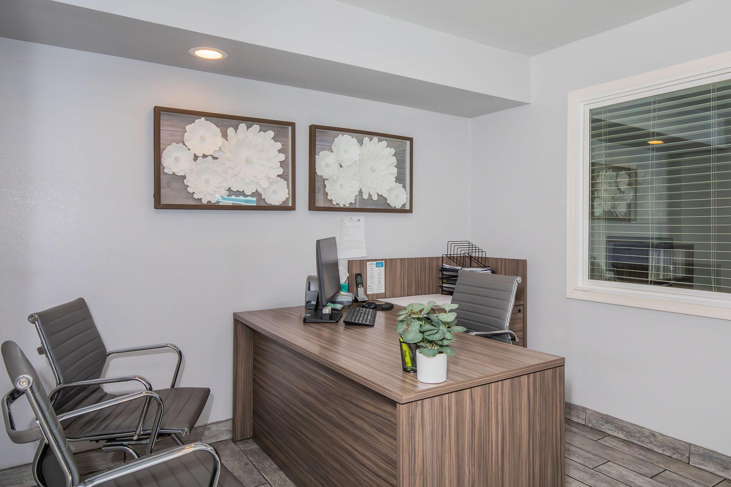 A modern office space featuring a wooden desk with a computer, a few office supplies, and a potted plant. Two sleek chairs are positioned in front of the desk. The light gray walls are decorated with two floral art prints, and a window with blinds is visible in the background.