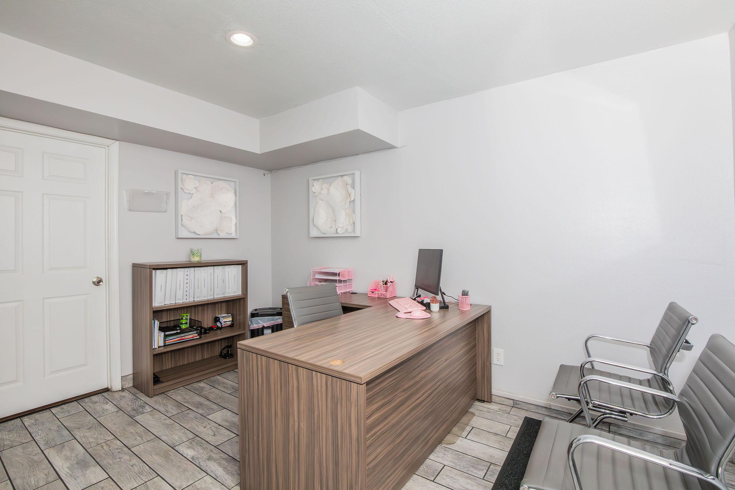 A modern office space with a wooden desk and a computer, featuring light gray walls and wood-style flooring. There are two metal chairs in front of the desk and decorative wall art. A closed door is visible in the background, along with a shelf displaying office items in the corner.
