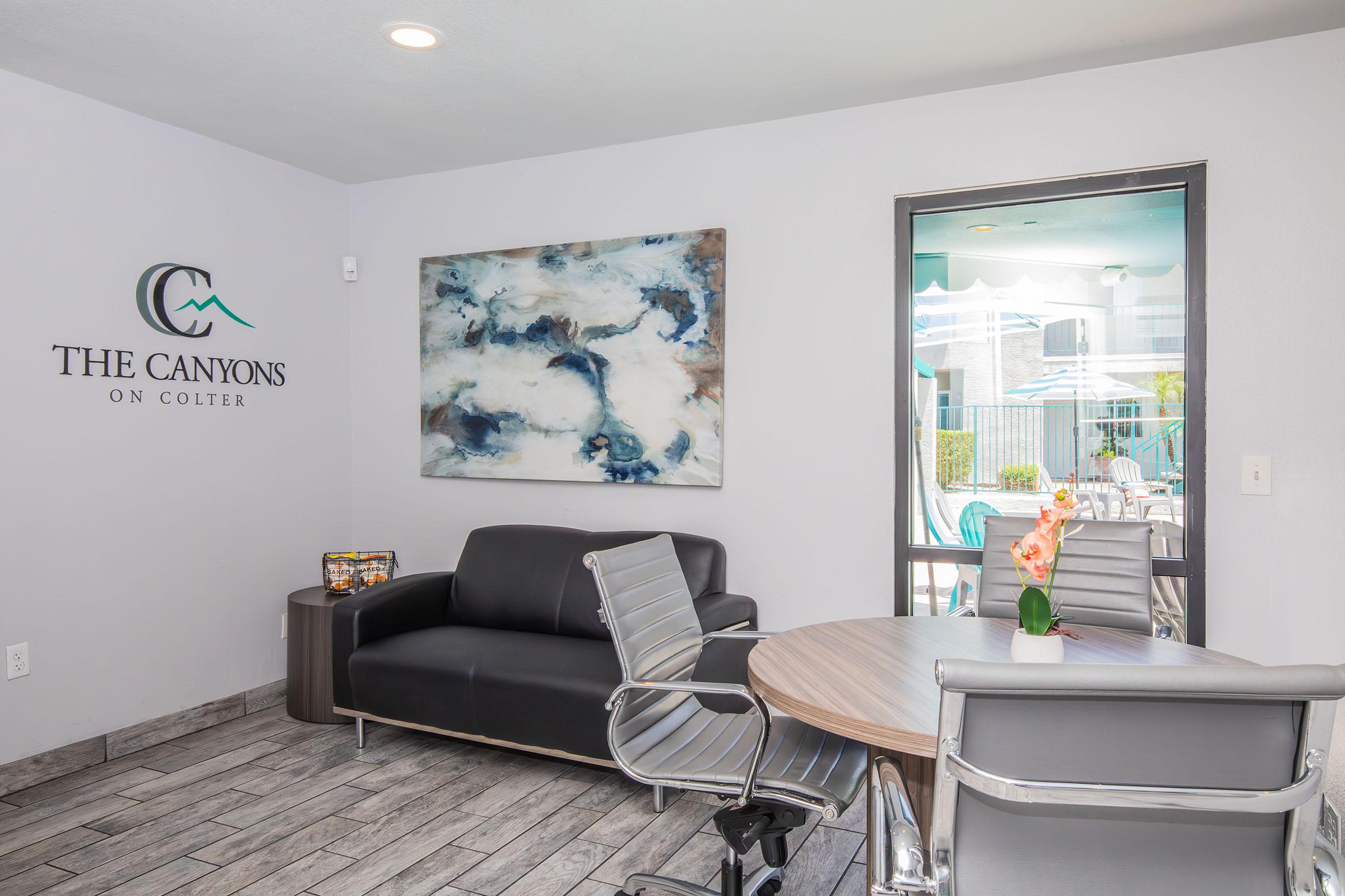 A modern lobby area featuring a black leather sofa, contemporary metal chairs, and a large abstract painting on the wall. There's a glass door leading to an outdoor pool area, with sunlight streaming in. The wall displays a logo reading "The Canyons on Colter." An indoor plant adds a touch of nature.