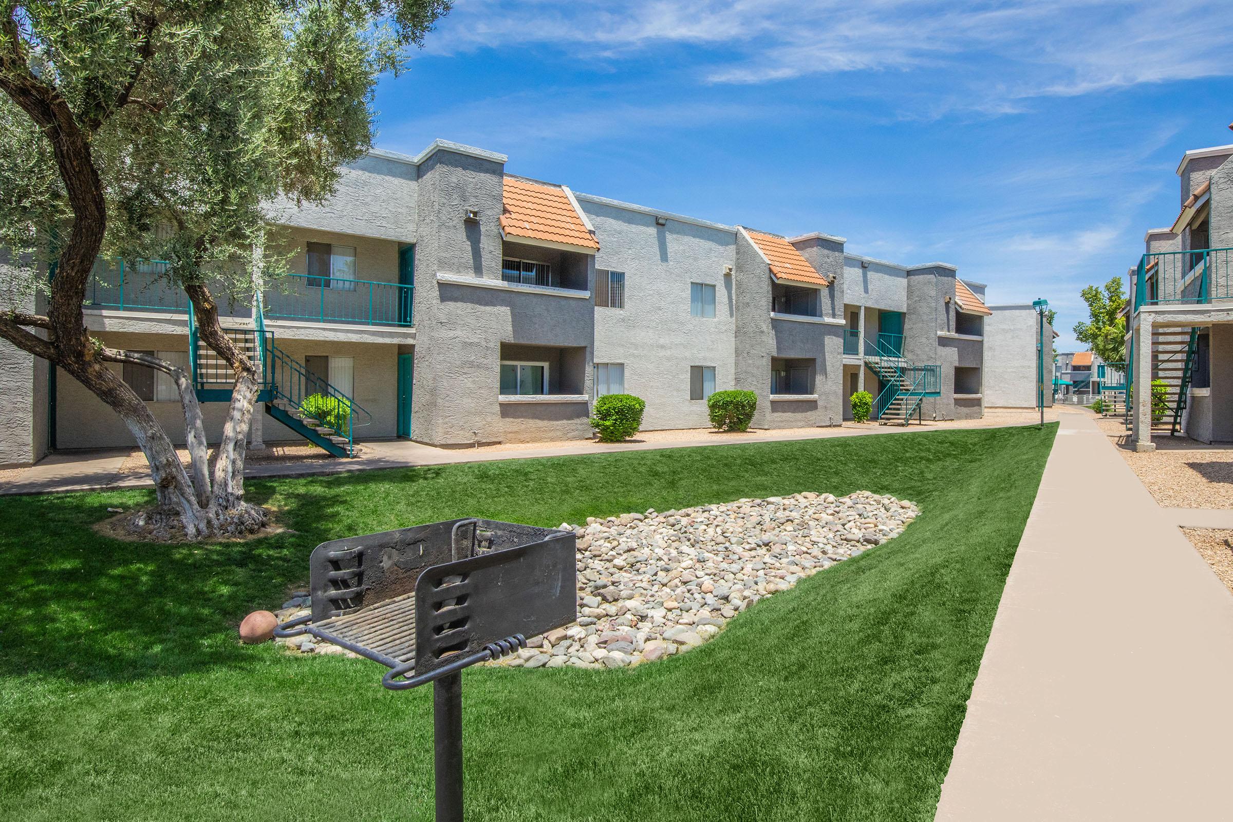 A view of a residential complex featuring two-story buildings with a gray exterior and orange rooftops, surrounded by neatly landscaped green grass and a rock garden. A walking path runs alongside the stone area, and trees provide shade in the outdoor space. The sky is clear and blue.