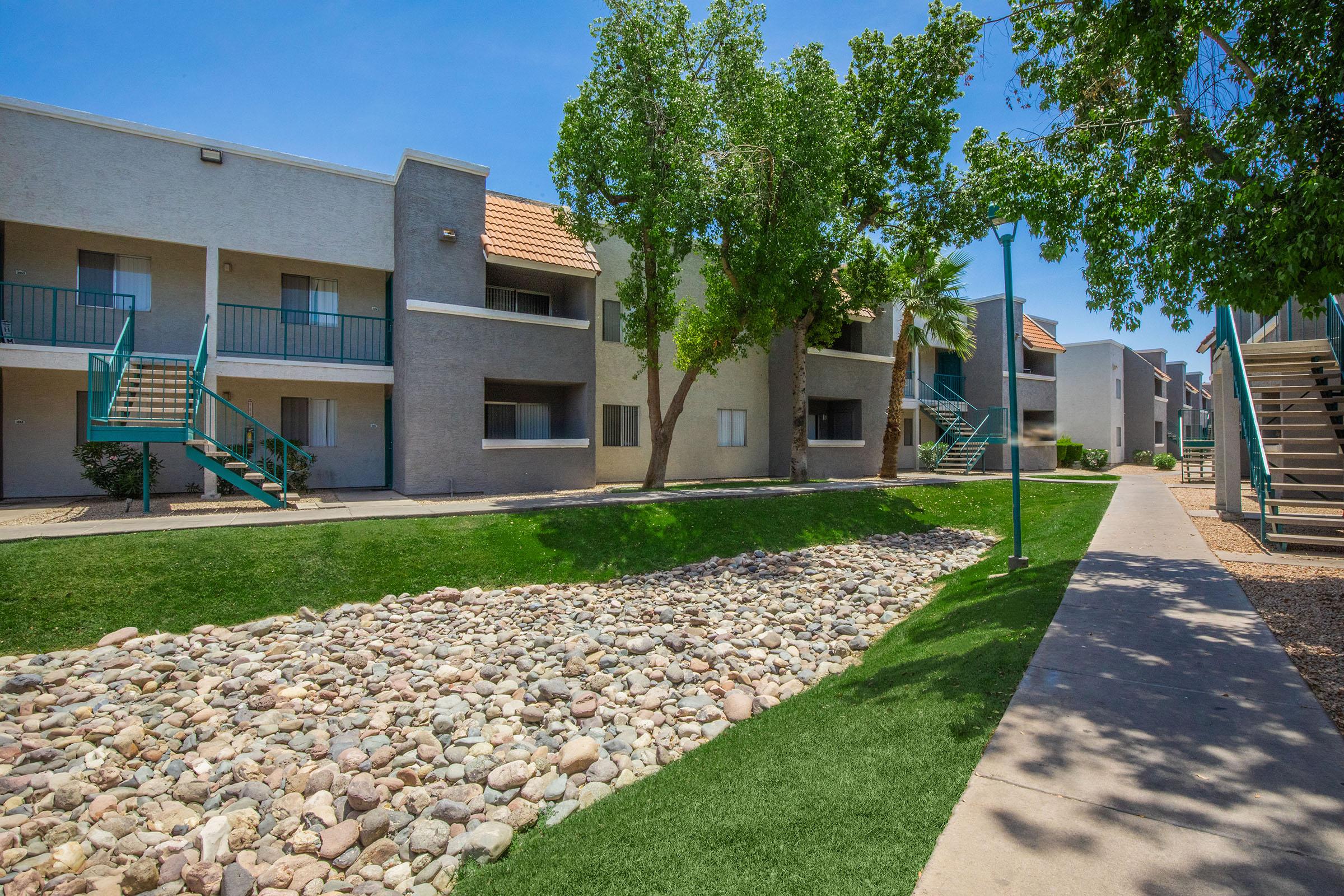 Apartment complex with two-story buildings, featuring green lawns and a pebble pathway. The buildings have balconies with stairs leading to the upper levels, and trees provide shade. A clear blue sky is visible above, creating a sunny and inviting atmosphere.