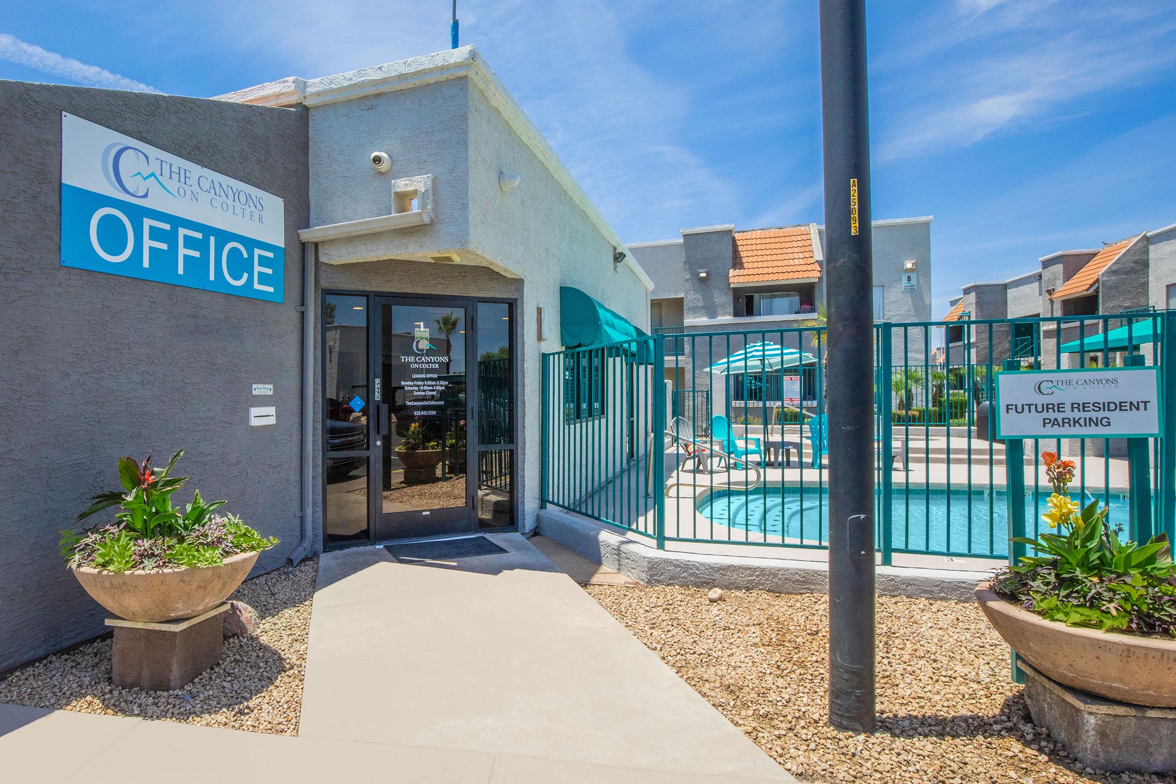 Office entrance of an apartment complex called "The Canyons on College," featuring a sign that says "OFFICE." A small swimming pool is visible in the background, surrounded by lounge chairs and umbrellas. The area has decorative plants and a gravel landscape, with a sign indicating "FUTURE RESIDENT PARKING."