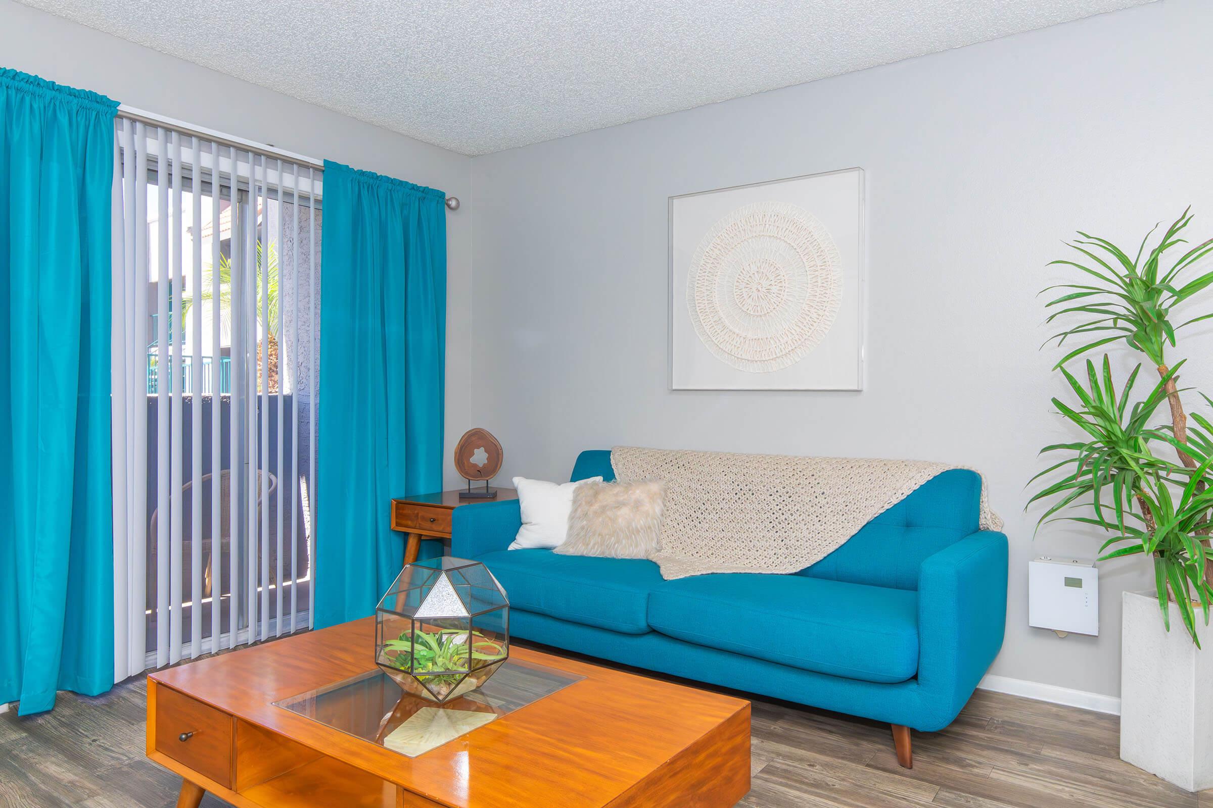 A bright and inviting living room featuring a turquoise couch adorned with a cozy blanket and decorative pillows. The space includes a wooden coffee table with a plant centerpiece, a large wall art piece, and vibrant blue curtains. A potted plant adds a touch of greenery to the room, enhancing its modern aesthetic.