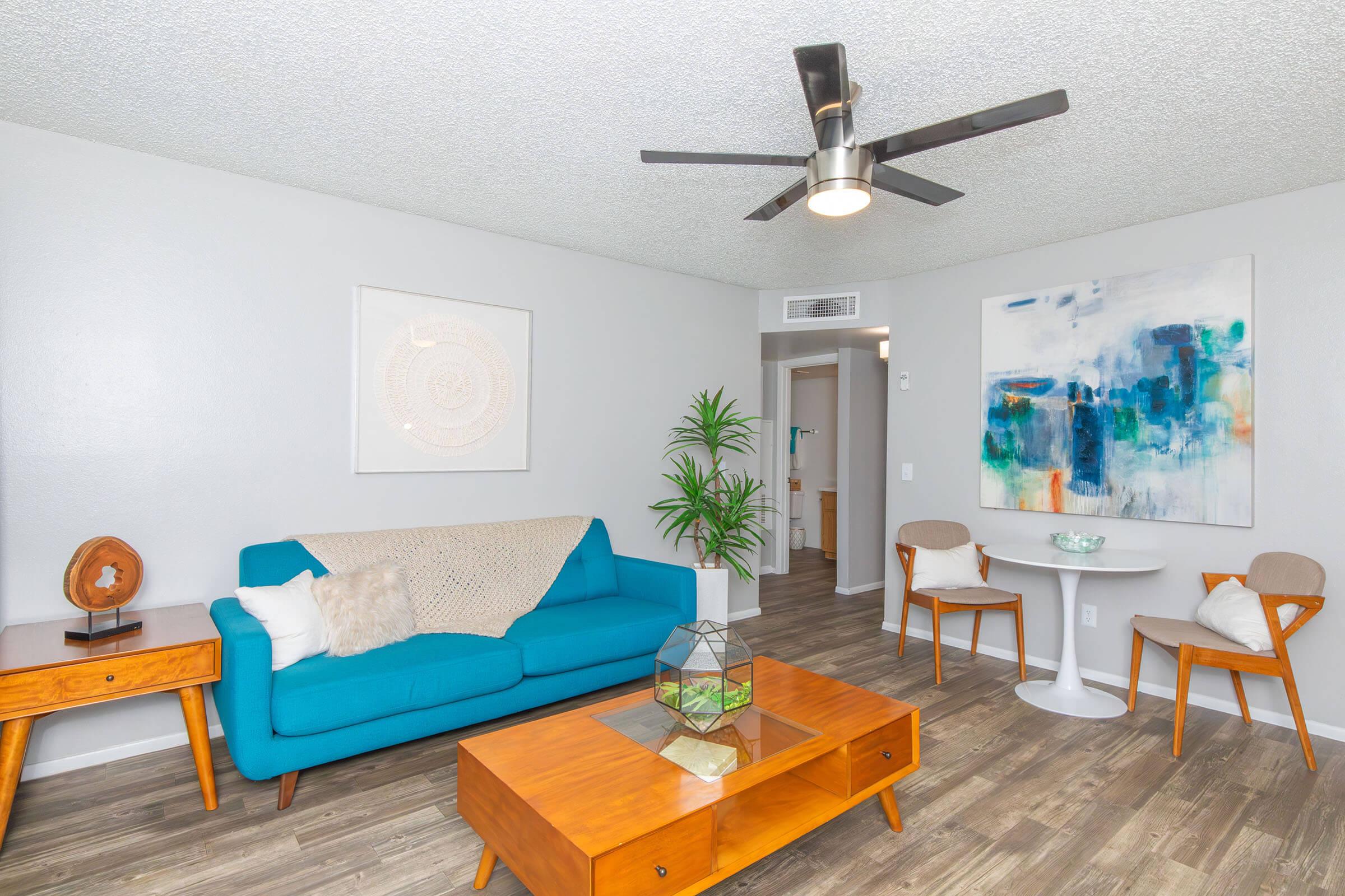 A modern living room featuring a bright blue couch, a wooden coffee table, and a decorative glass terrarium. The walls are adorned with abstract art, and there is a small dining area with a round table and stylish chairs. A potted plant adds a touch of greenery, and a ceiling fan provides a contemporary feel.