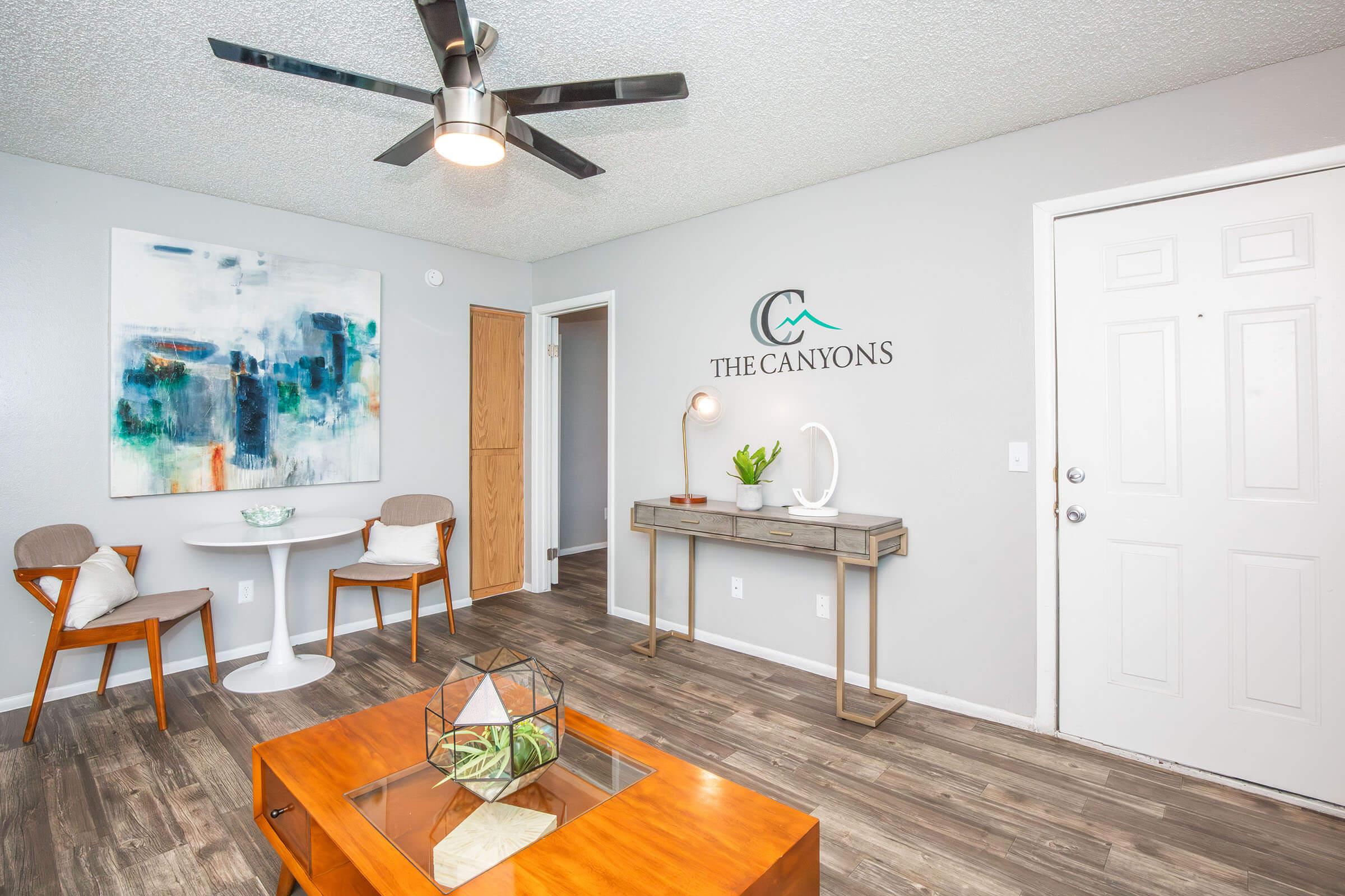 A cozy living space featuring a light gray wall, modern ceiling fan, and stylish furnishings. There is a round white dining table with two wooden chairs, a wooden console table displaying a decorative lamp and a geometric terrarium, and a door leading to another room. The Canyons logo is displayed on the wall.