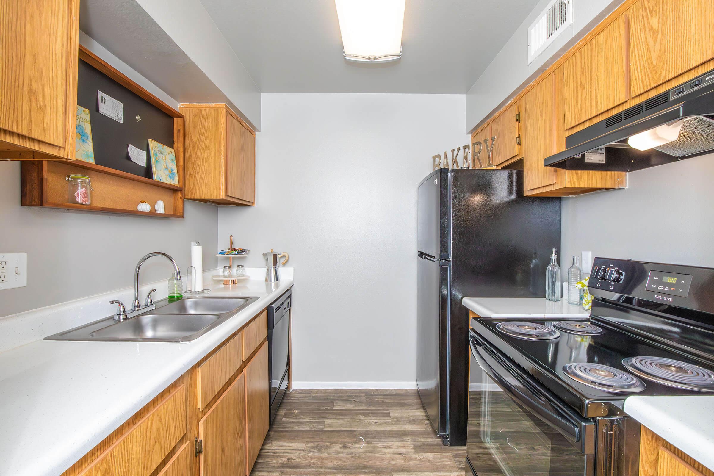 A modern kitchen with wooden cabinets, stainless steel sink, and black appliances. The image features a stove, oven, and refrigerator against a light gray wall. Decorative items are placed on the shelves, and the countertop is clean and minimalist. Soft lighting illuminates the space, creating a cozy atmosphere.