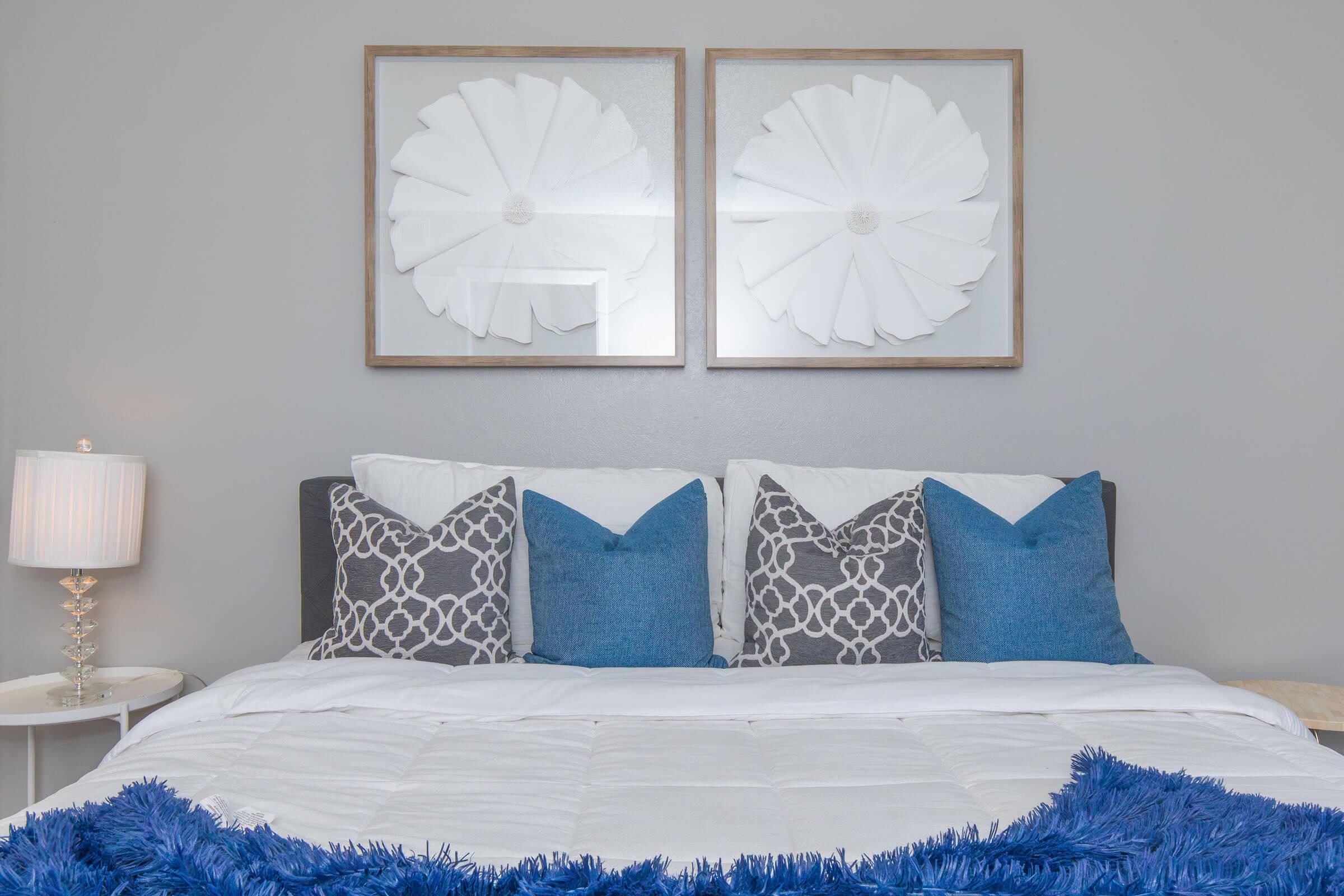 A cozy bedroom featuring a neatly made bed with white bedding and decorative pillows in gray and blue. Above the bed, two framed wall art pieces resembling large white flowers add a stylish touch. A bedside lamp provides soft lighting, enhancing the inviting atmosphere of the room.