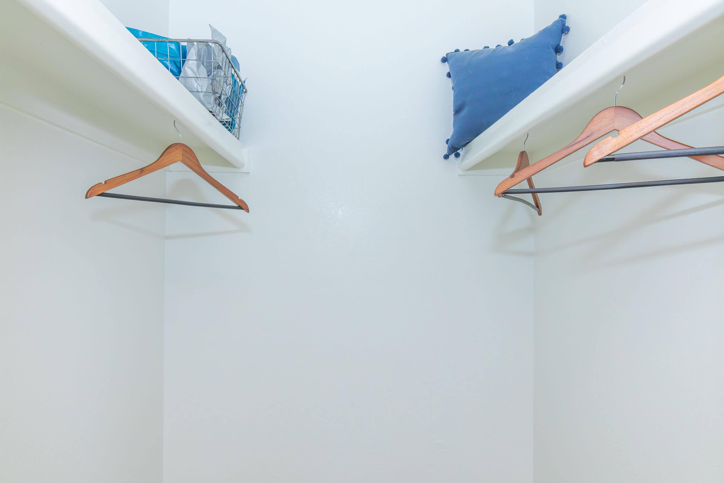 Empty closet with two wooden hangers on the left and one on the right. A blue pillow sits on a shelf, and a wire basket is visible on the left shelf. The walls are white, creating a clean and minimalist look.