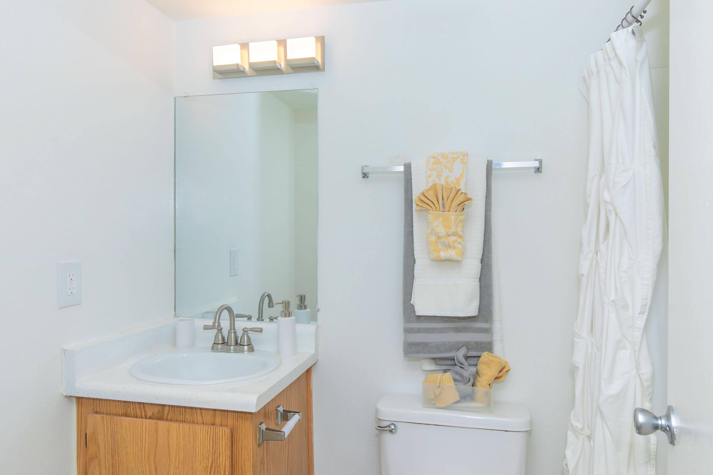 A clean, modern bathroom featuring a white sink with silver faucet, a mirror above the sink, and a light fixture. Towels are neatly hung on a towel bar, with a decorative towel displayed. A white shower curtain is partially visible, and a toilet is seen with rolled towels beside it. The walls are painted white, creating a bright atmosphere.
