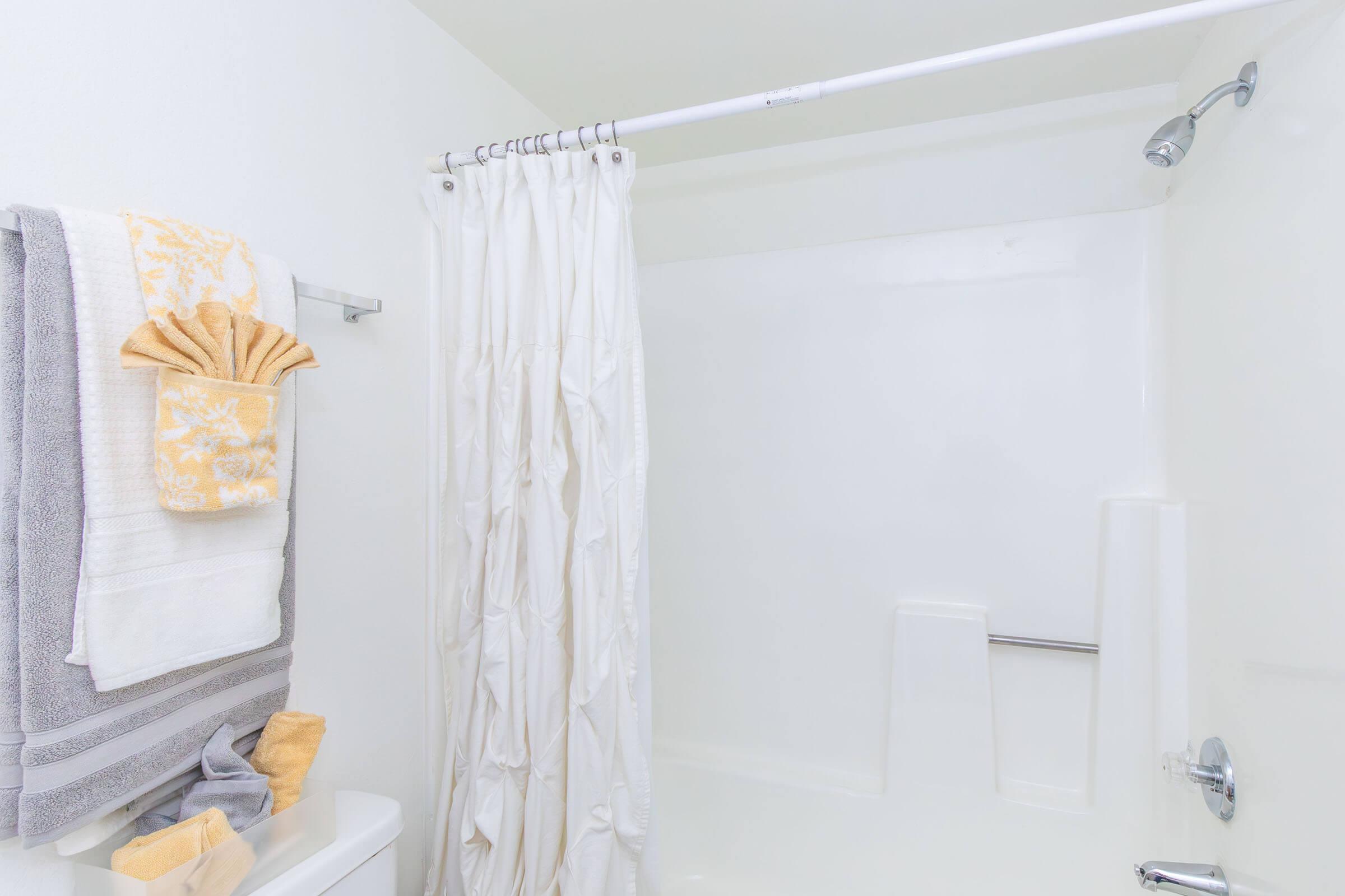 A clean, minimalistic bathroom with a white shower curtain and light-colored towels hanging on a towel rack. The shower area has white walls and a simple bathtub design, creating a bright and modern ambiance.