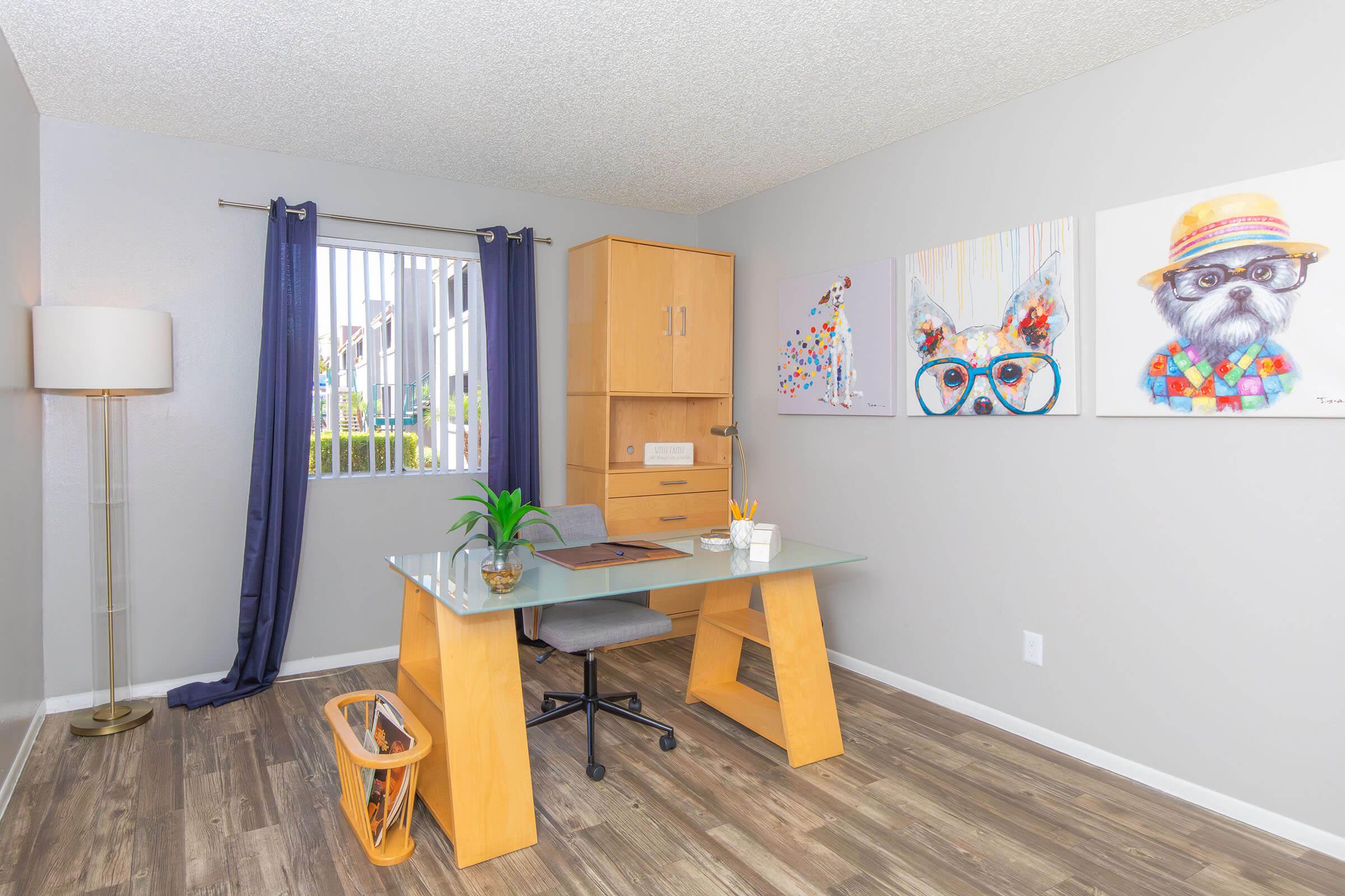 A modern office space featuring a glass desk with a wooden base, a comfortable chair, and a small plant. The walls are decorated with colorful animal paintings and there is a tall lamp in one corner. Natural light filters through a window with blue curtains, creating a bright and inviting atmosphere.