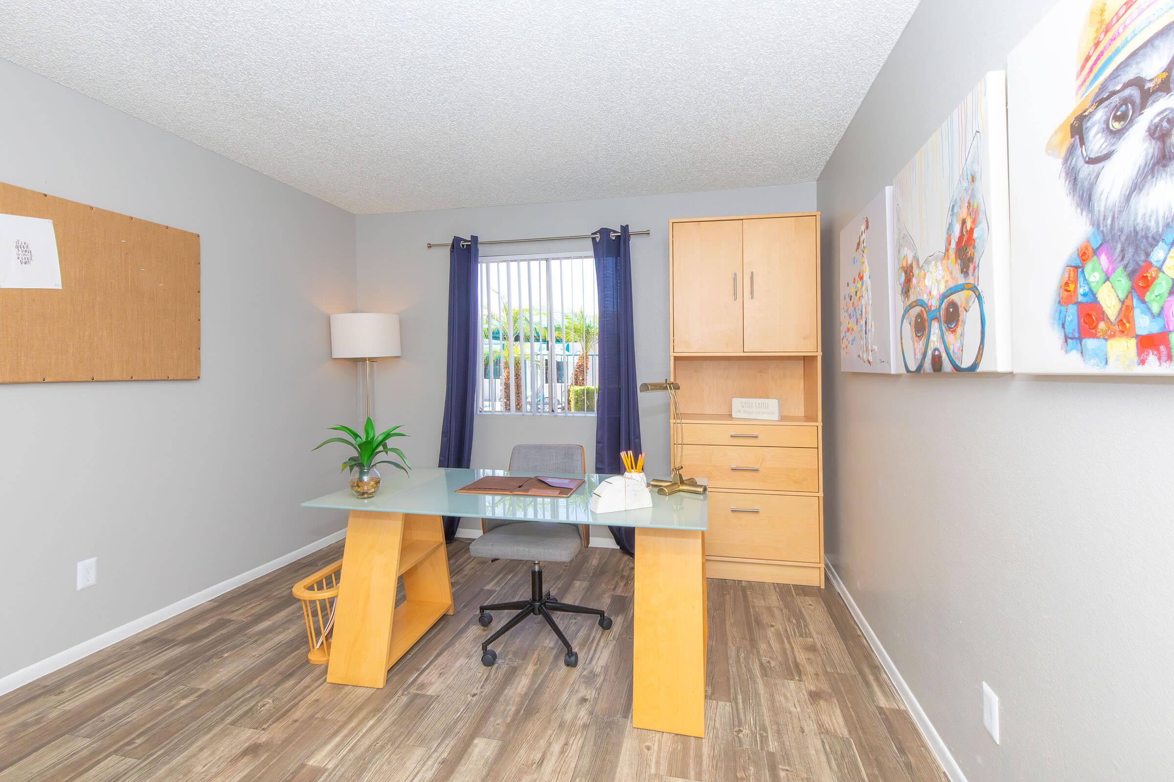 A modern office space featuring a glass desk with a gray chair, a lamp, and a potted plant. The walls are light gray with a corkboard and colorful artwork. A wooden storage unit stands beside a window covered with blue curtains, allowing natural light into the room. The floor is covered with light wood laminate.