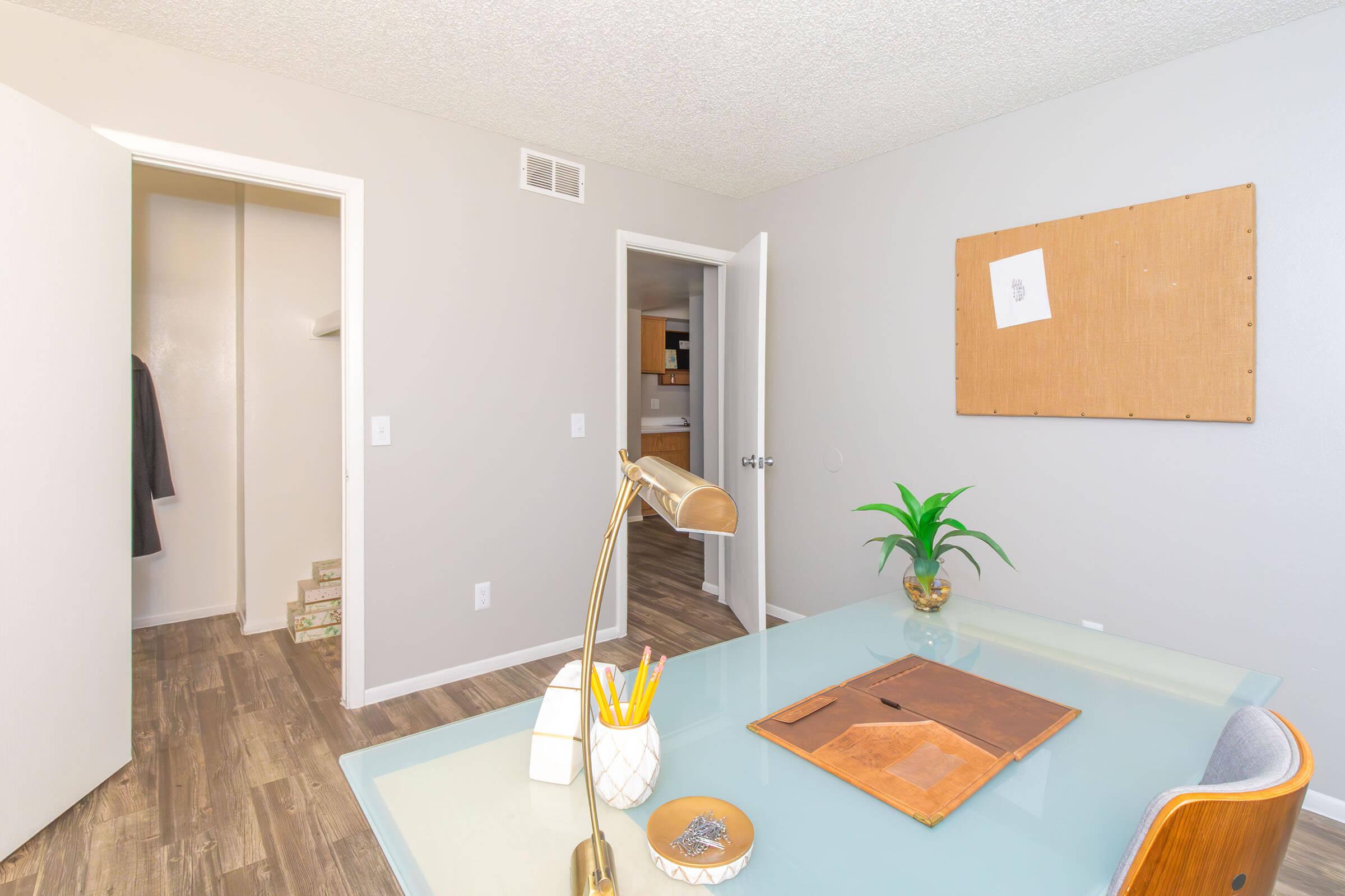 A modern home office with a glass desk, a stylish lamp, and decorative items. Two doors lead to adjacent rooms, while a large corkboard is mounted on the wall. The floor is made of laminate wood, and a plant adds a touch of greenery to the workspace. Soft, neutral colors create a calm atmosphere.