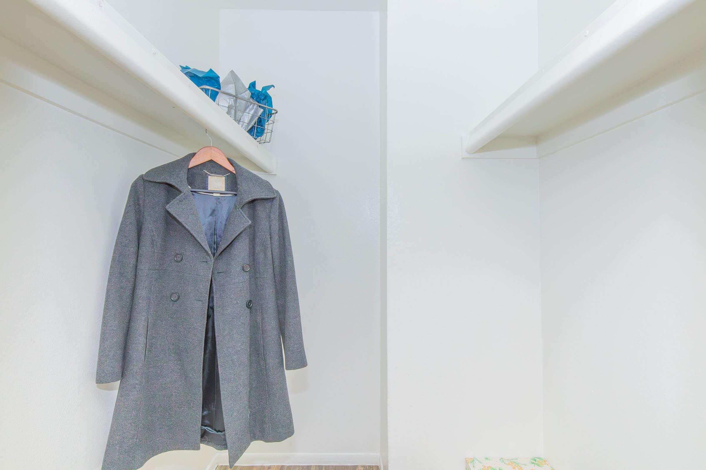 A gray coat hanging on a wooden hanger inside a sparse closet. The closet has white walls and two shelves with some blue bags and a basket on the top shelf. The floor is clear, creating a minimal and uncluttered appearance.