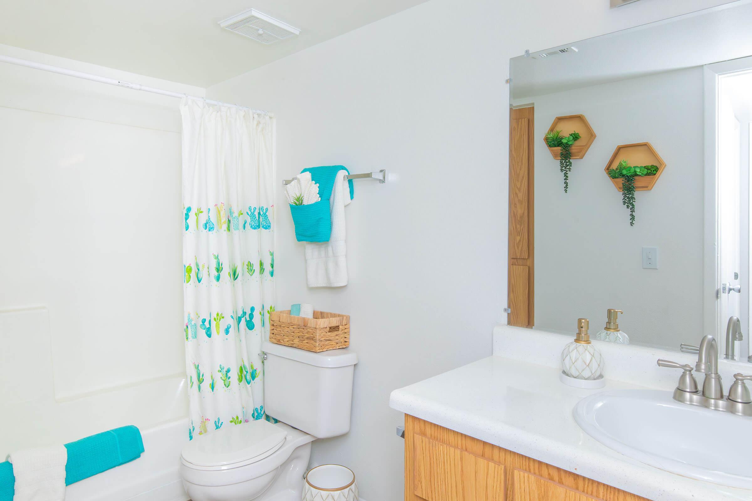 Bright and airy bathroom featuring a white shower curtain with a colorful cactus pattern, a beige basket on the countertop, and greenery in decorative hexagonal wall shelves. The sink area includes a round mirror, modern faucets, and a clean, minimalist design with light-colored cabinetry.