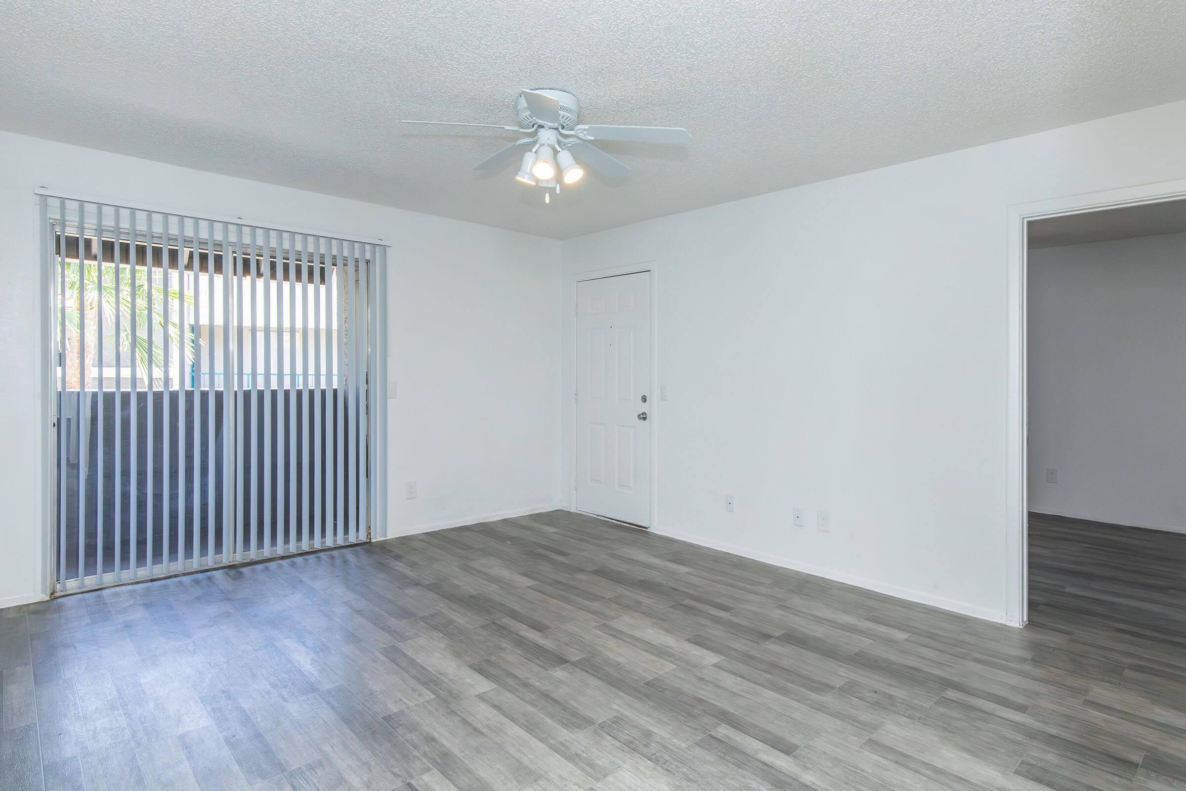 A bright and empty living room featuring light-colored walls and a wood-look floor. There is a ceiling fan with lights, a window with vertical blinds letting in natural light, and a door leading to another room. The room appears spacious and ready for furnishing.