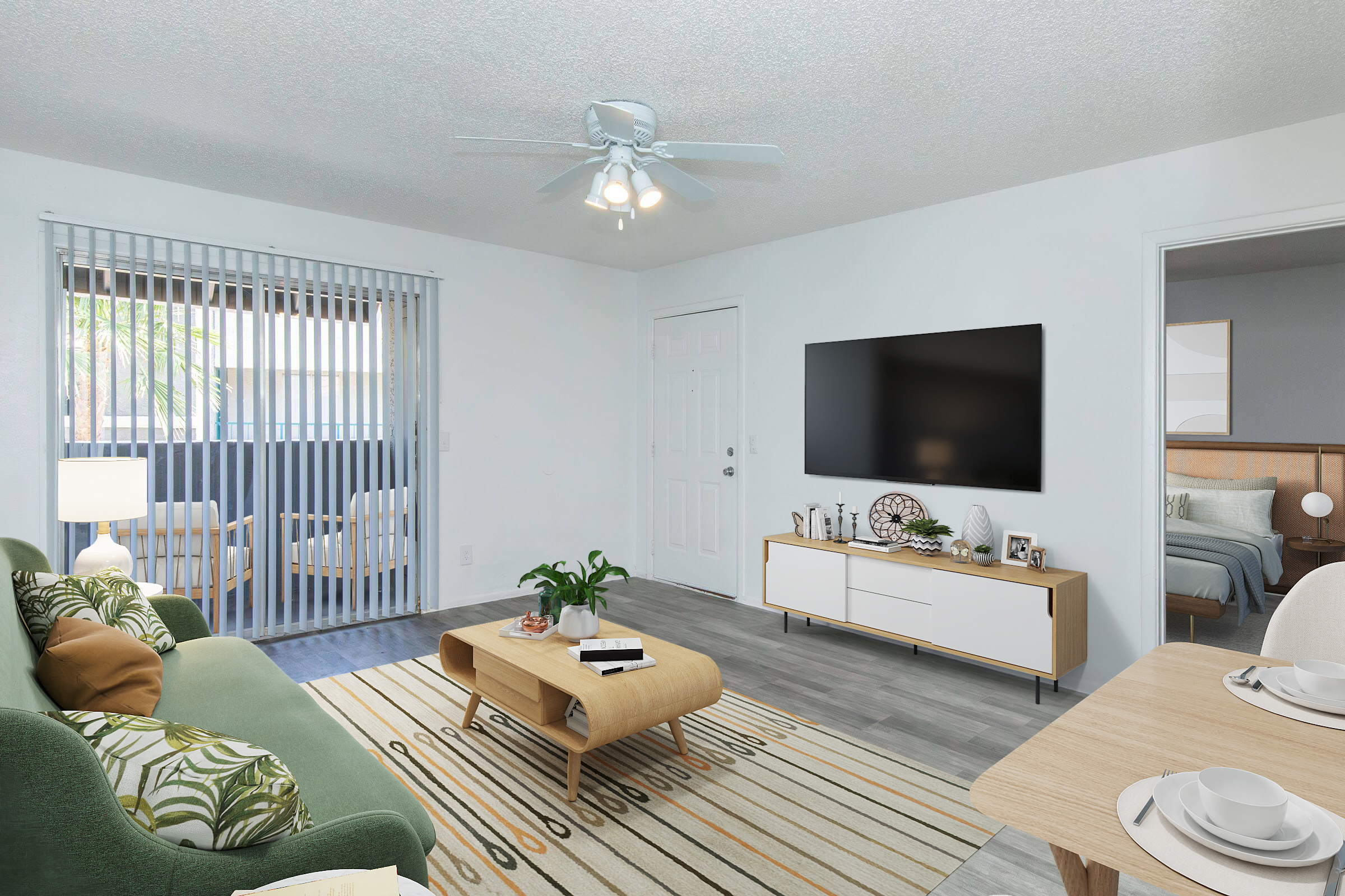A modern living room featuring a green sofa with patterned cushions, a light wooden coffee table, and a flat-screen TV mounted on the wall. The space has a sliding glass door leading outside, light-colored walls, and a cozy dining area with a beige table setting. A potted plant adds a touch of greenery.